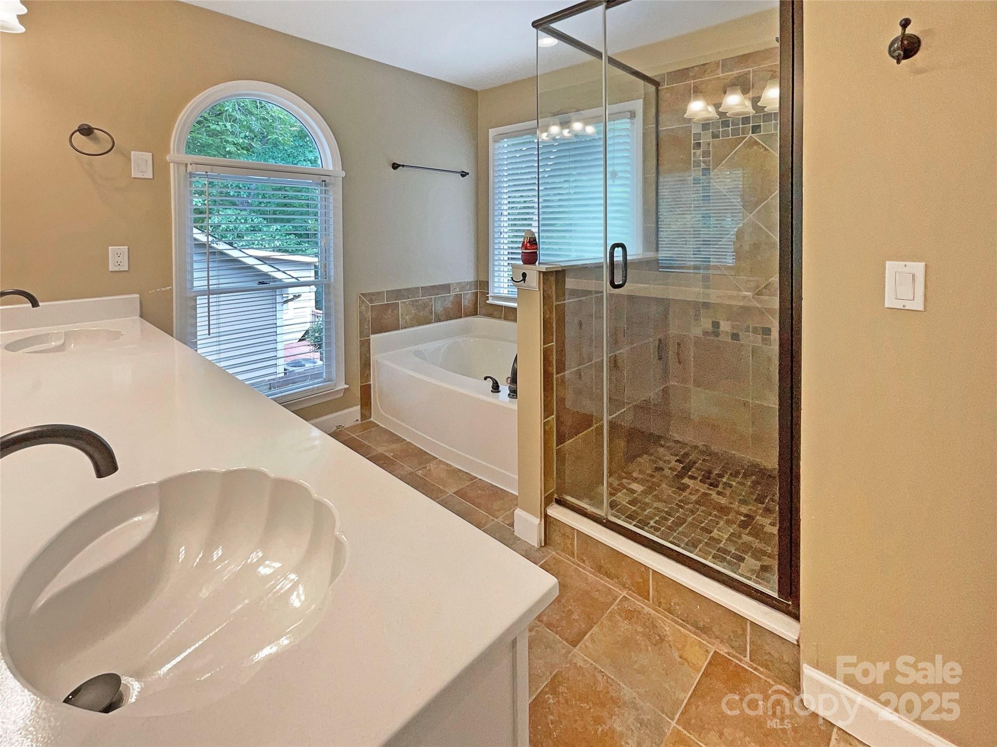 Bathroom with a double vanity, arched window over the tub, and glass-enclosed tiled shower.