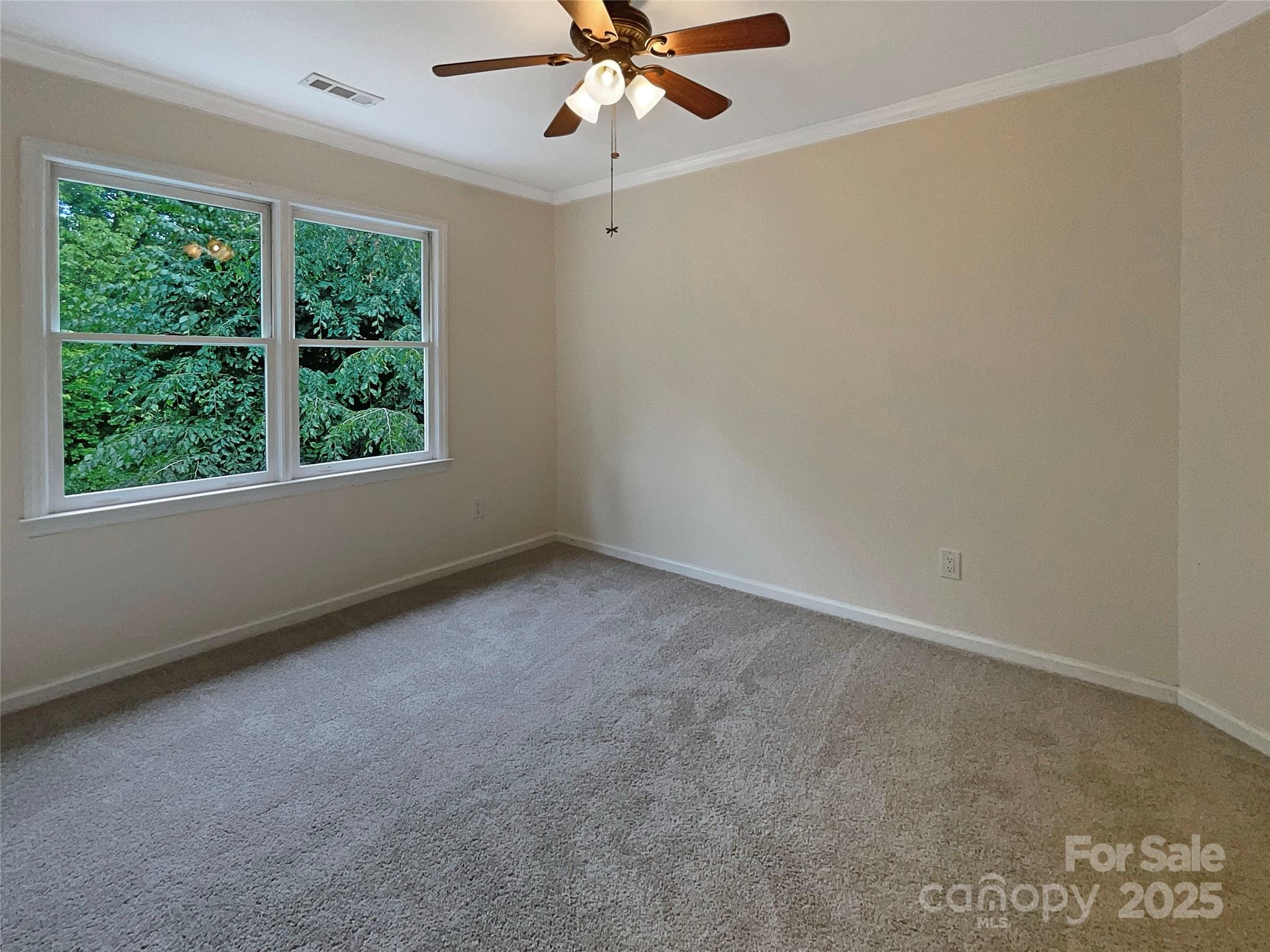 Empty carpeted bedroom with two windows overlooking dense greenery and a ceiling fan.