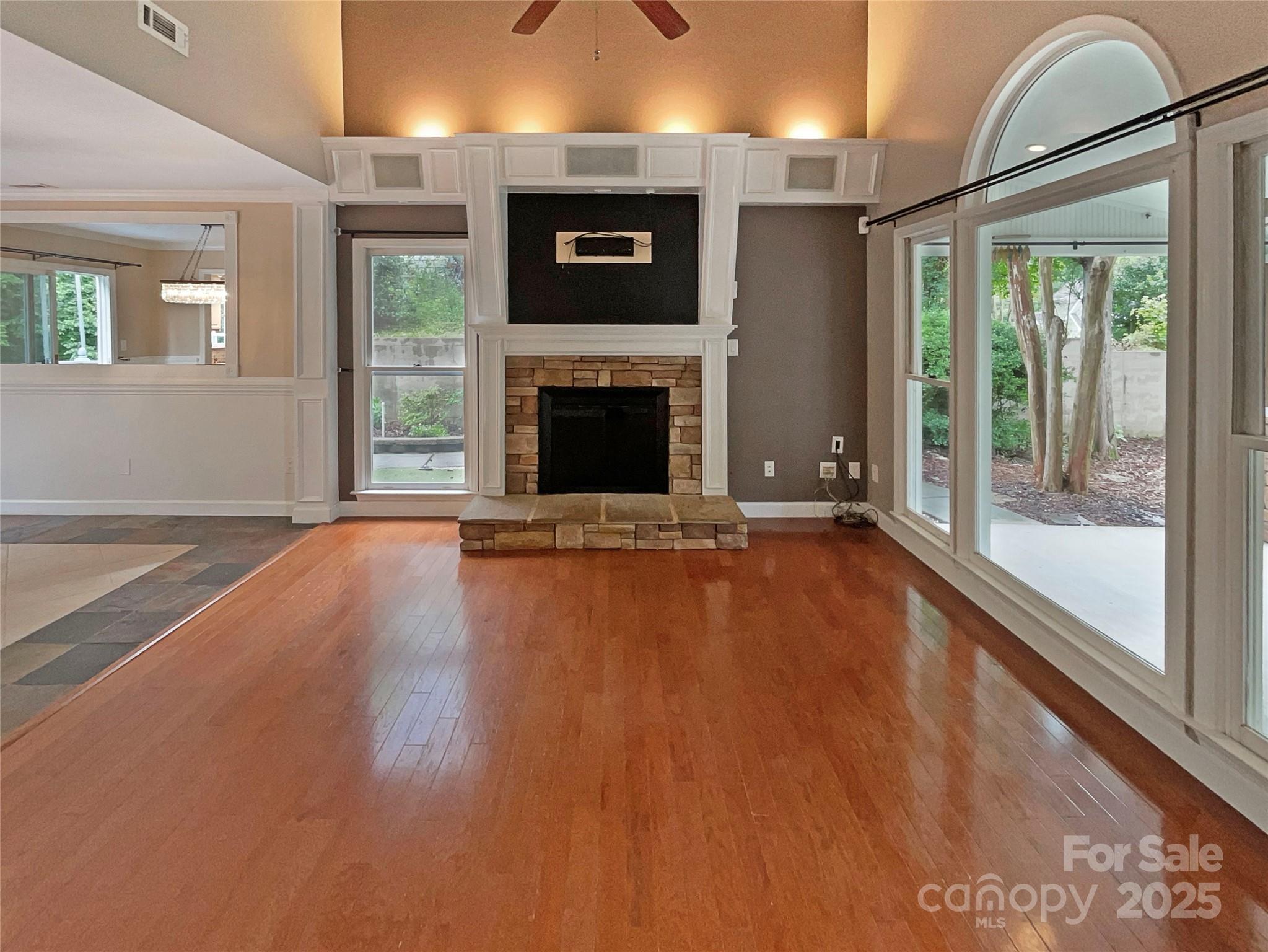Vacant living room with hardwood floors, a stone fireplace, and large windows overlooking a wooded yard.
