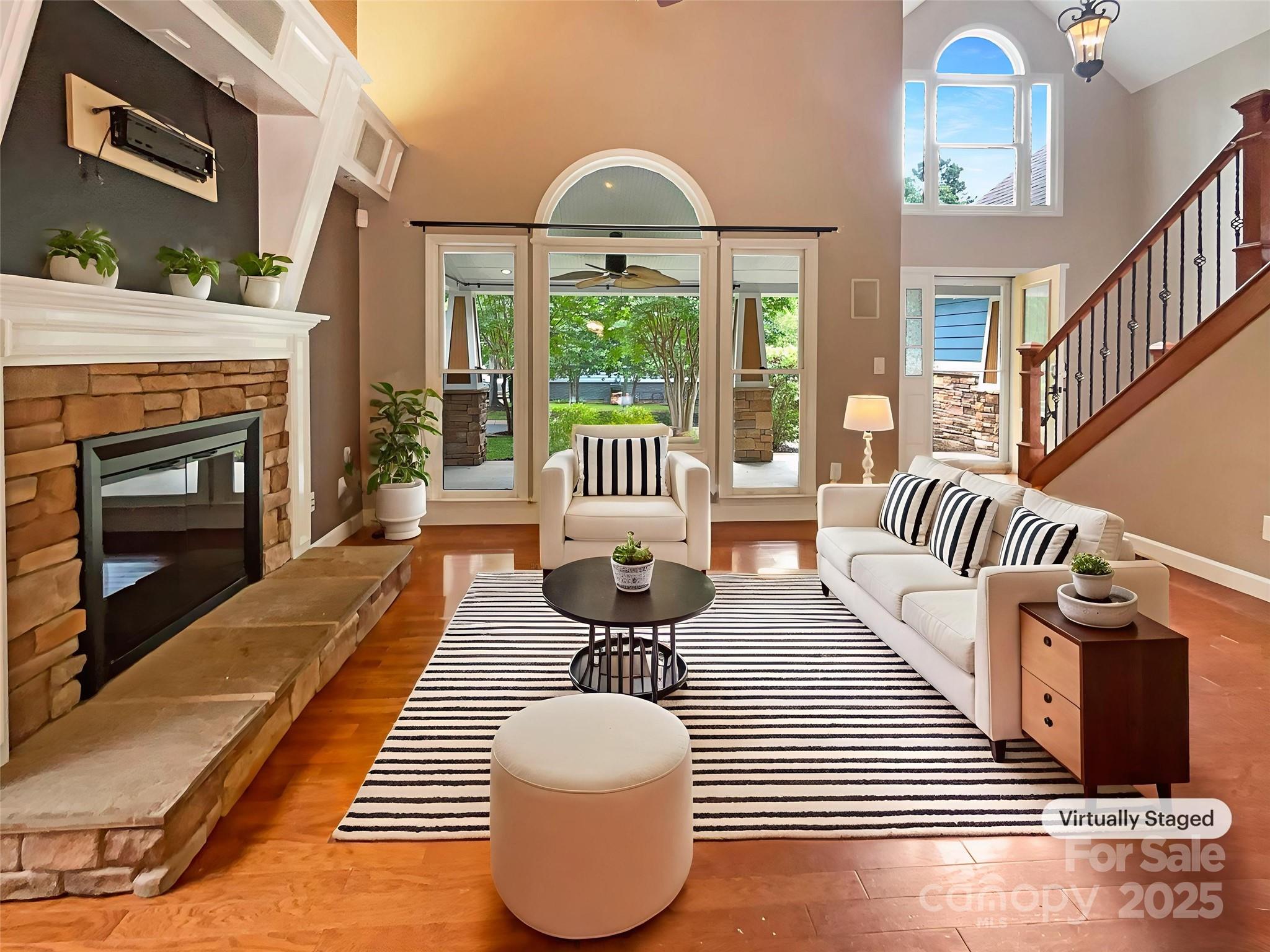 Living room with a stone fireplace, white seating, striped rug, and staircase.