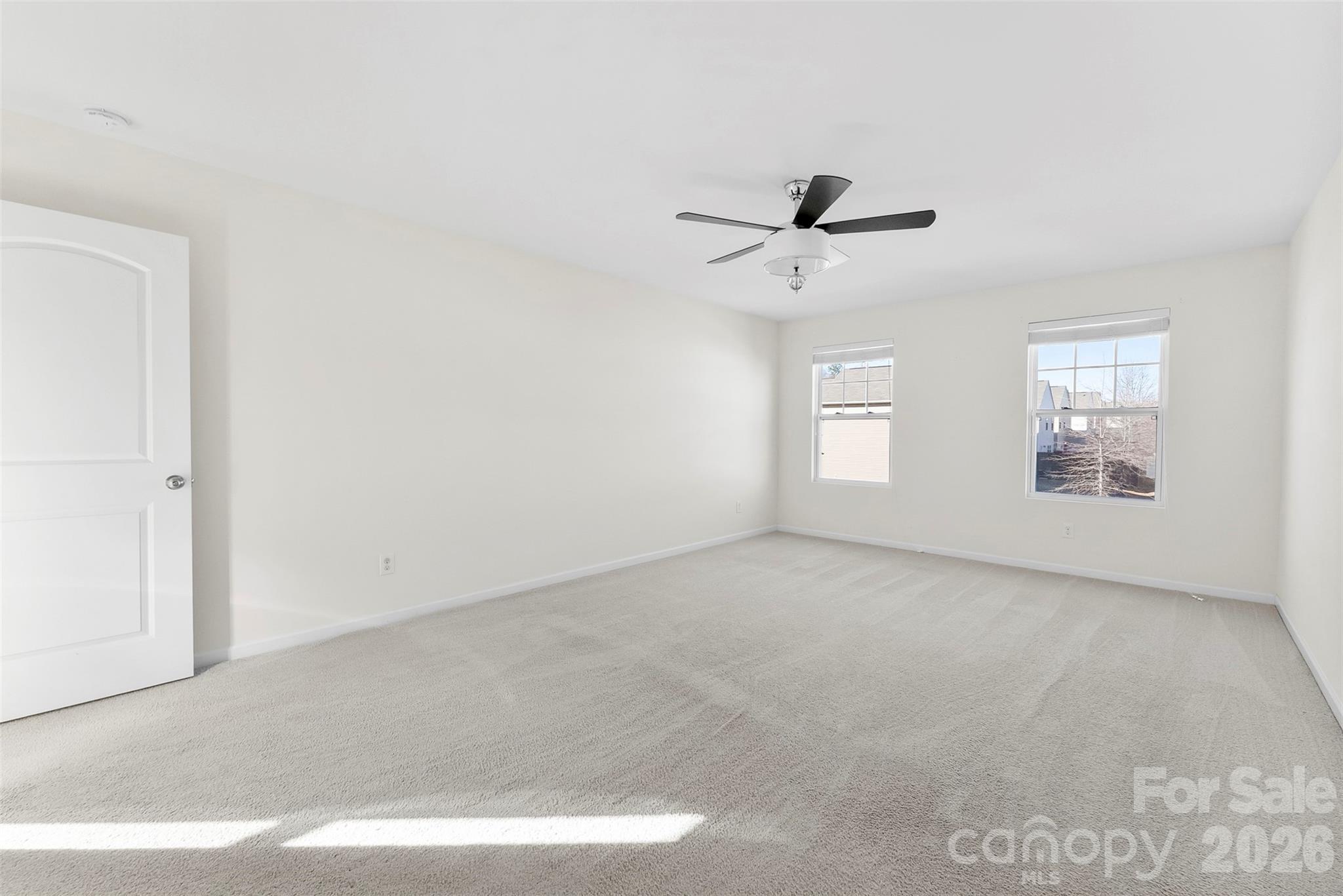 Empty bedroom with light carpet, a dark-bladed ceiling fan, and two sunlit windows.