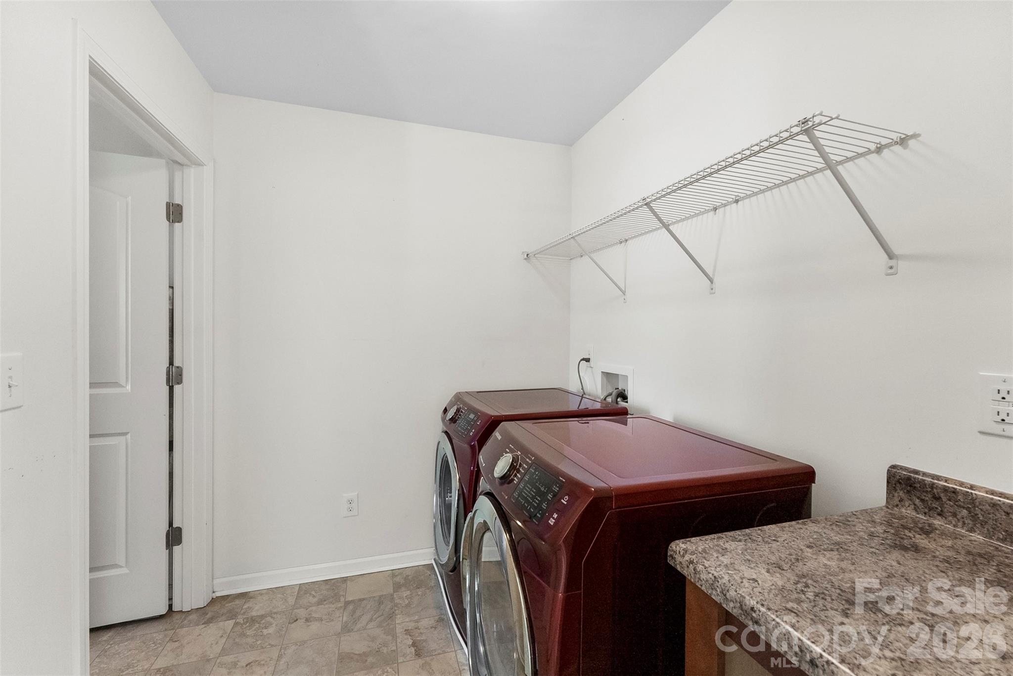 Laundry room with burgundy washer and dryer, overhead wire shelf, and speckled countertop.