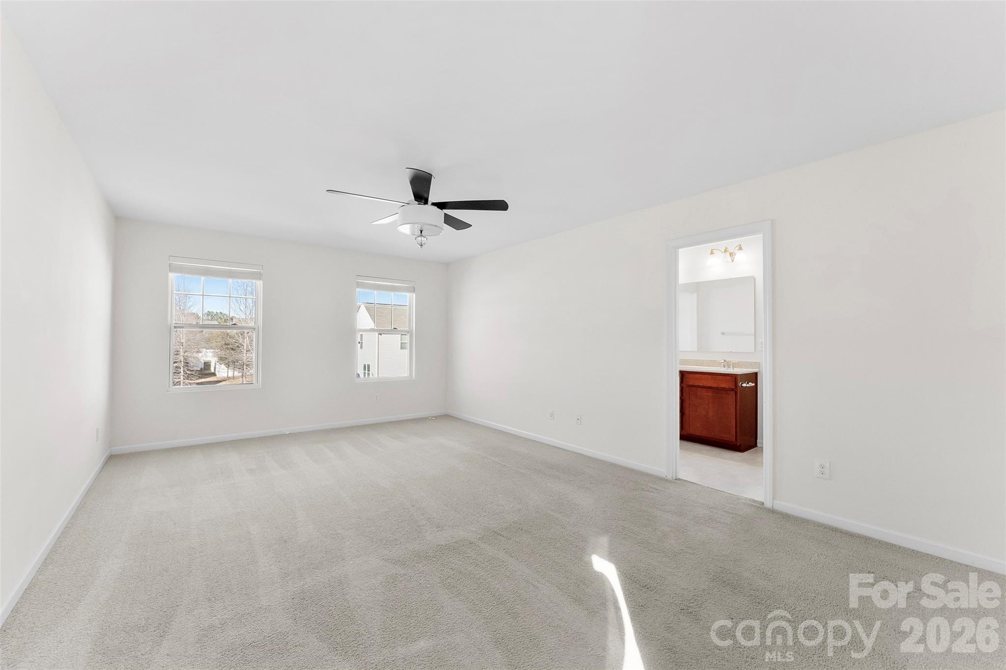 Empty carpeted bedroom with two windows, a black ceiling fan, and an adjoining bathroom vanity.