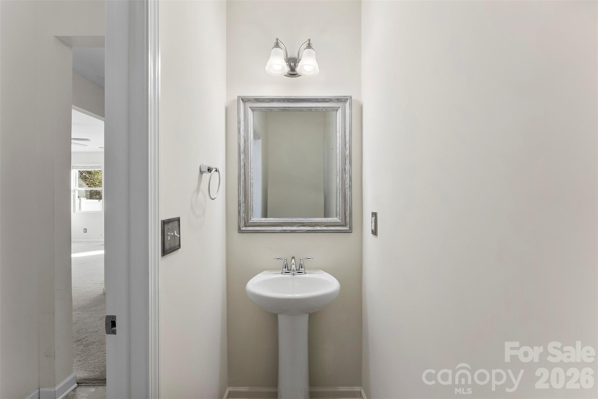Bathroom with a white pedestal sink, framed mirror, and two-light fixture beside an open doorway.