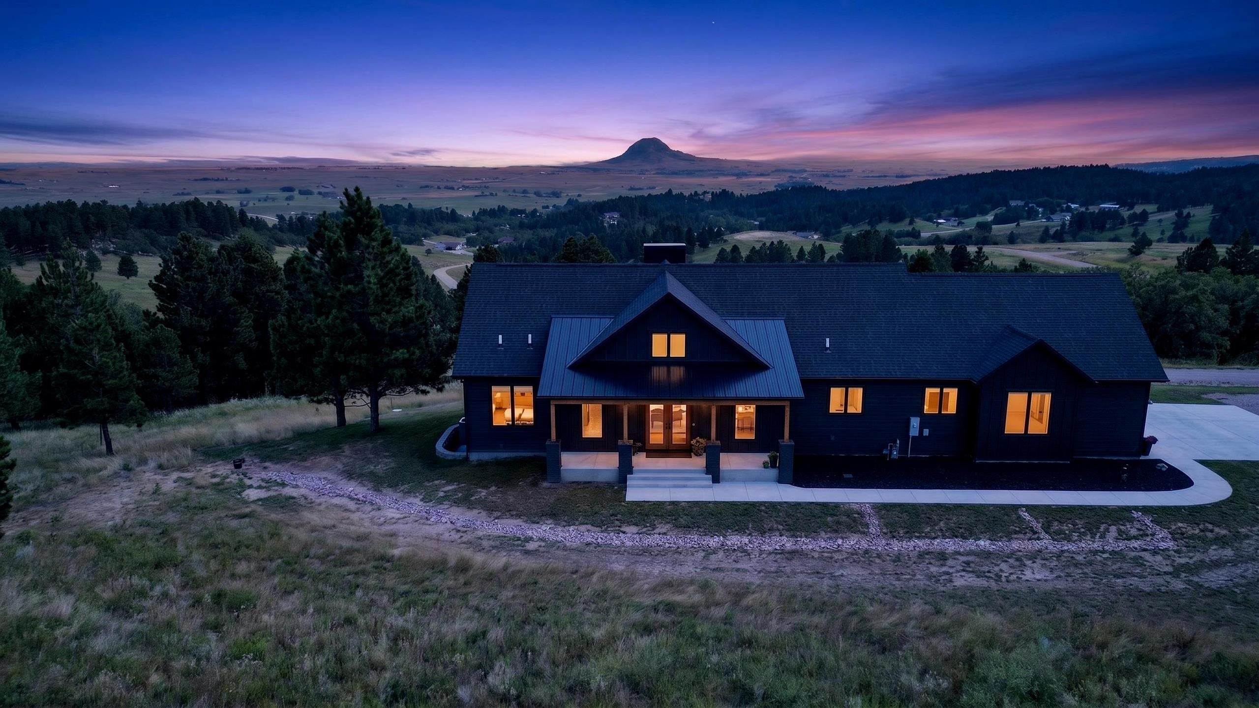 Modern dark house with glowing windows at dusk, framed by pine trees and a rounded hill on the horizon.