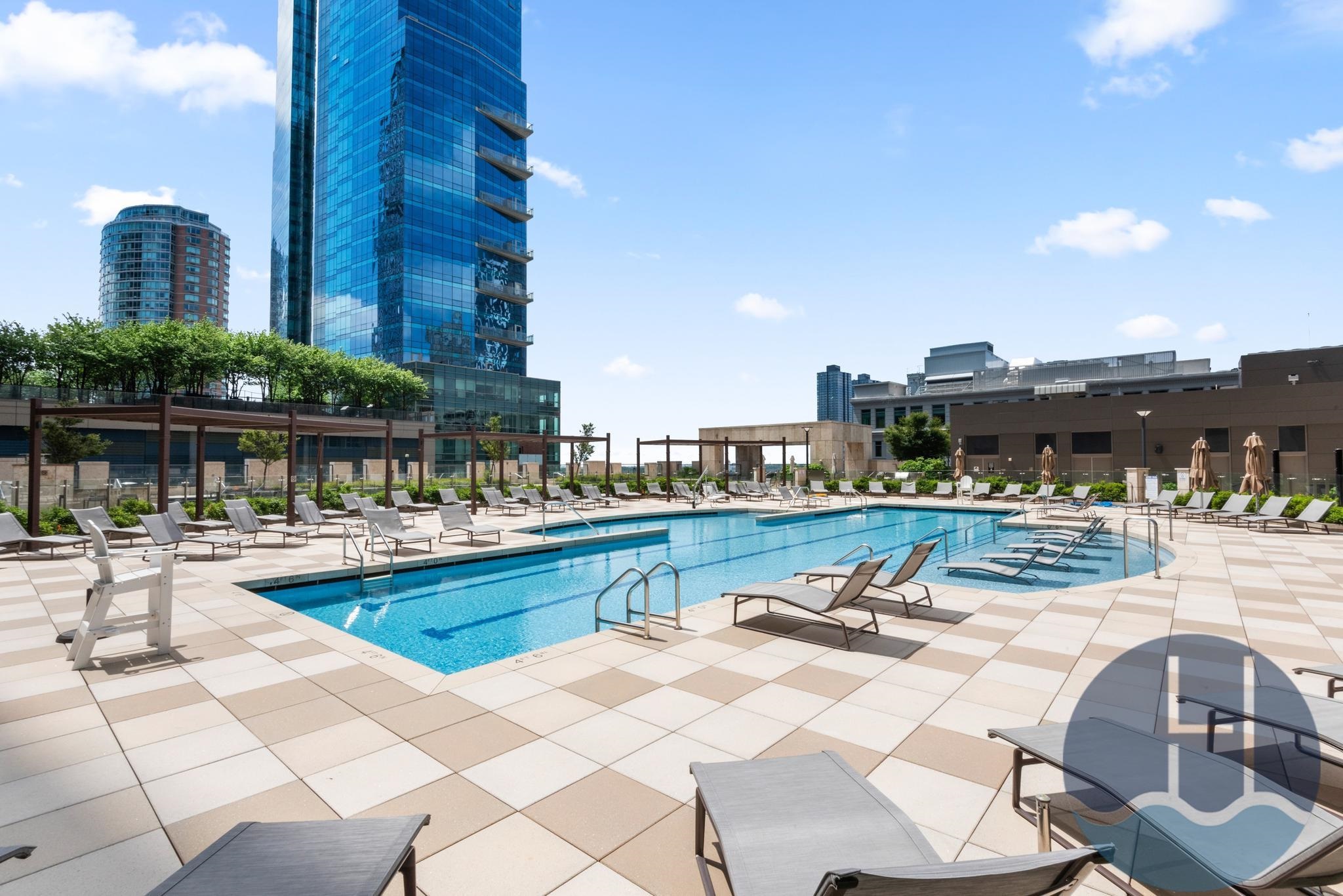Rooftop pool surrounded by lounge chairs and a tiled sun deck, with city towers behind it.