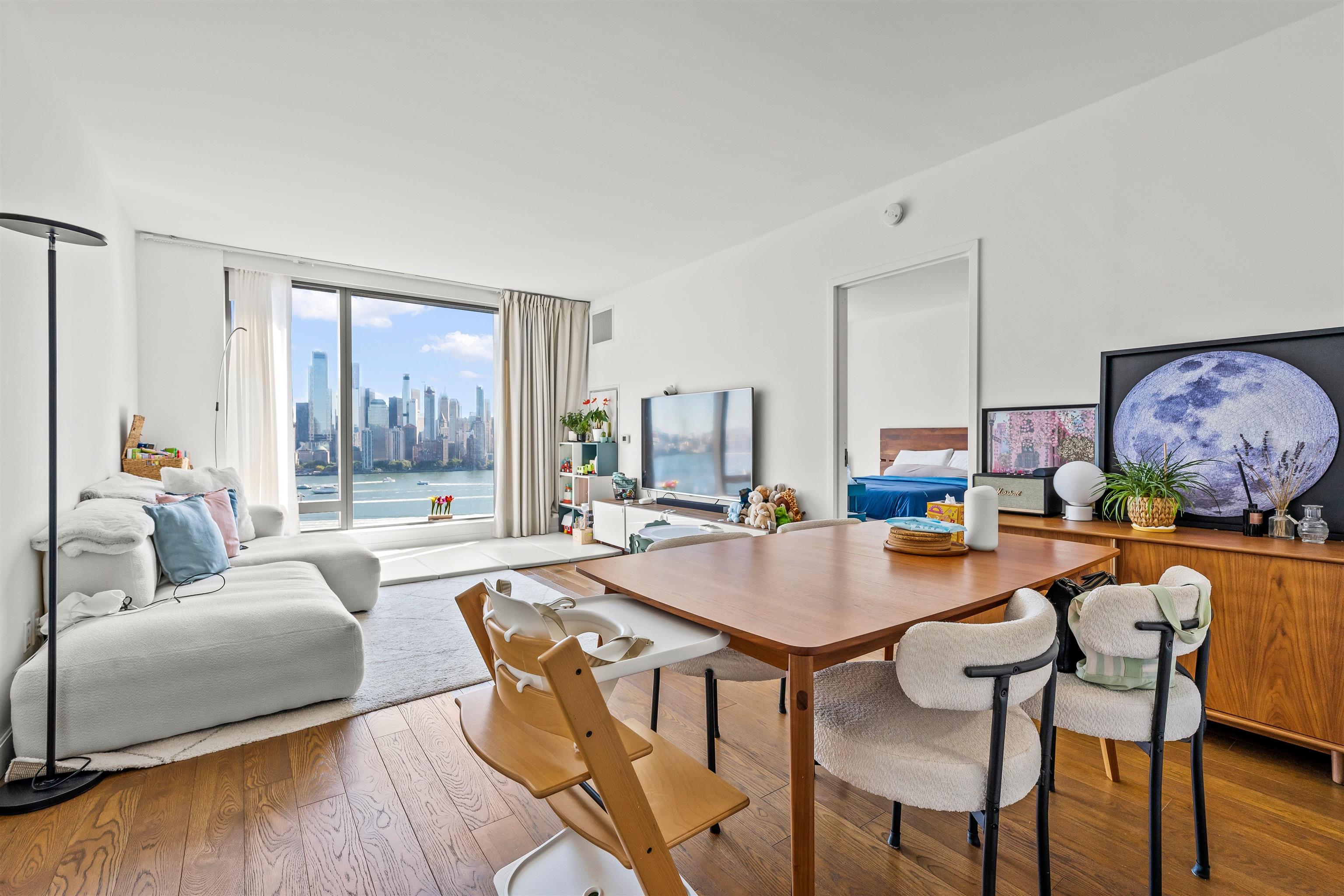 Furnished living and dining area with floor-to-ceiling windows overlooking the water and Manhattan skyline.