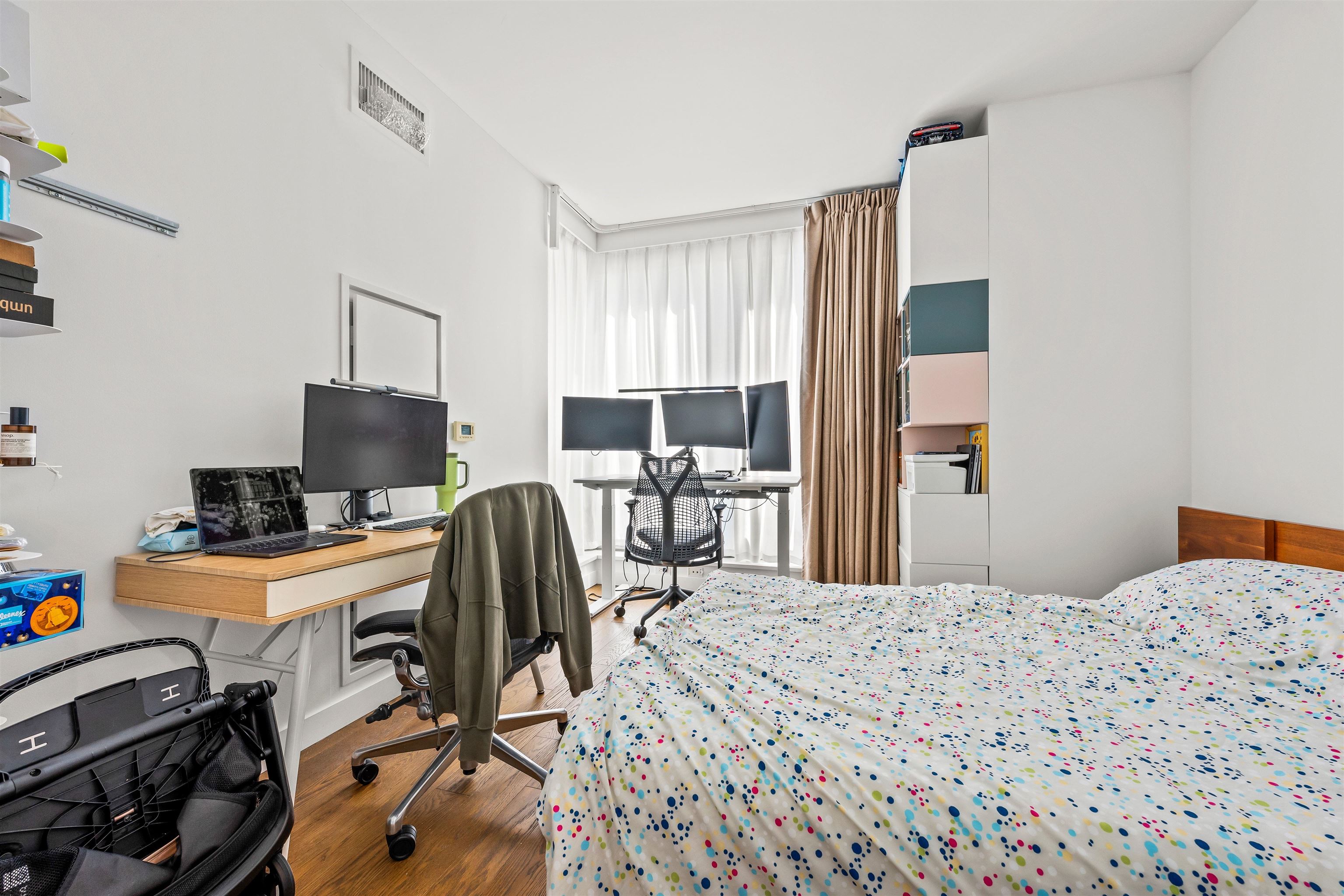 Bedroom with dotted bedding and two computer workstations beside bright curtained windows.