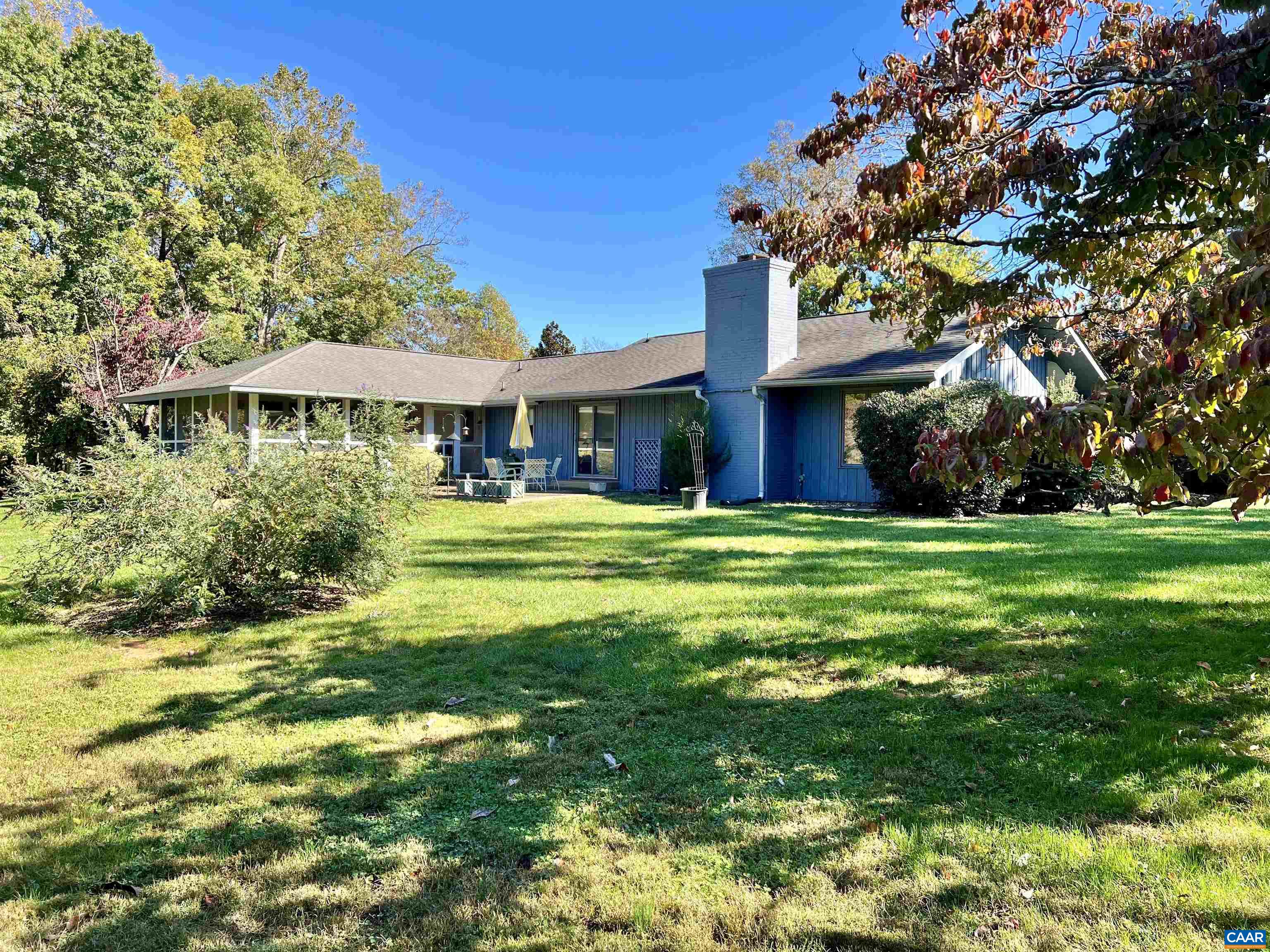 Blue single-story home with a chimney, patio seating, and a tree-shaded lawn.