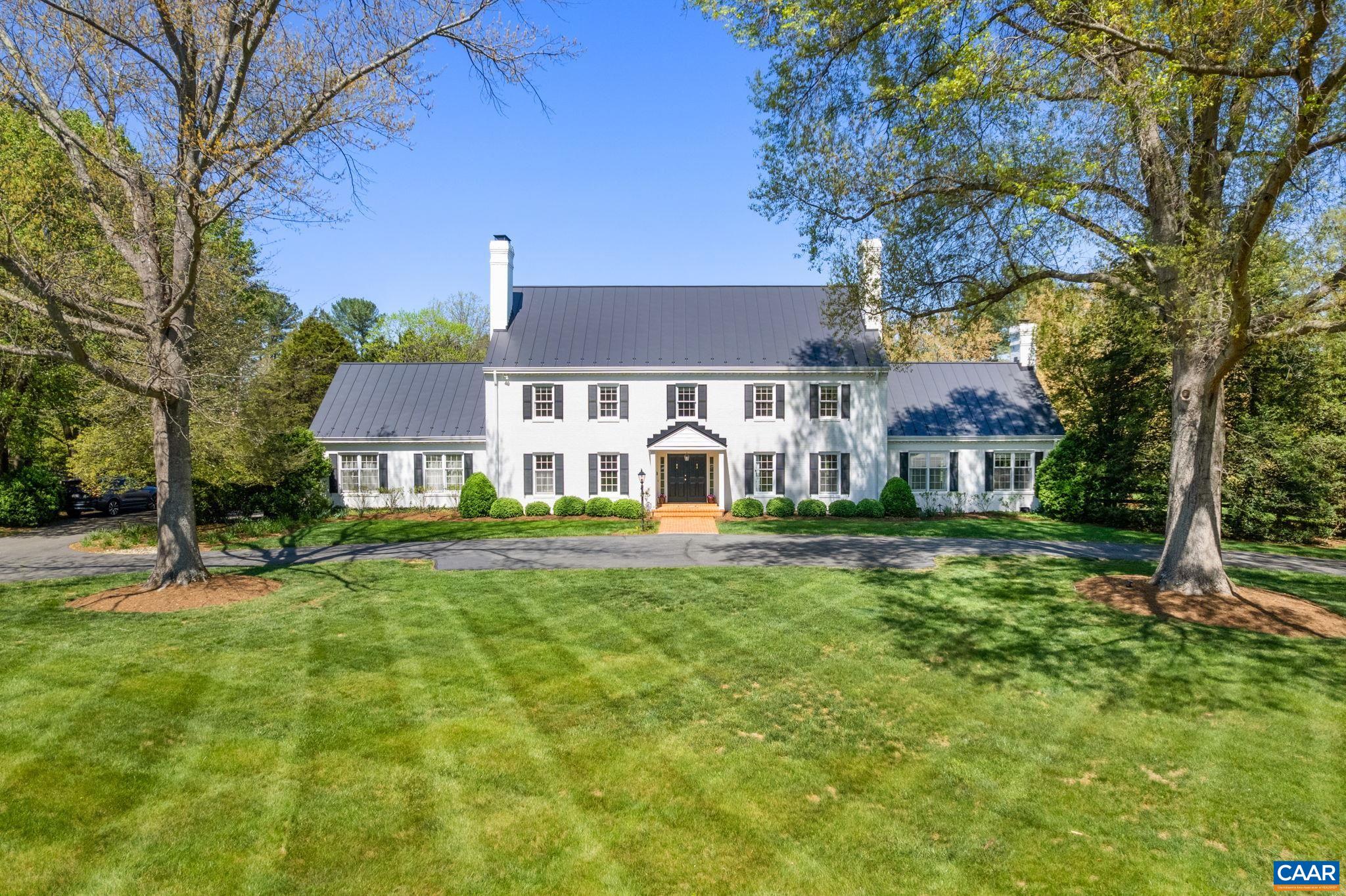White Colonial-style house with black shutters and a dark metal roof, framed by mature trees and a broad lawn.