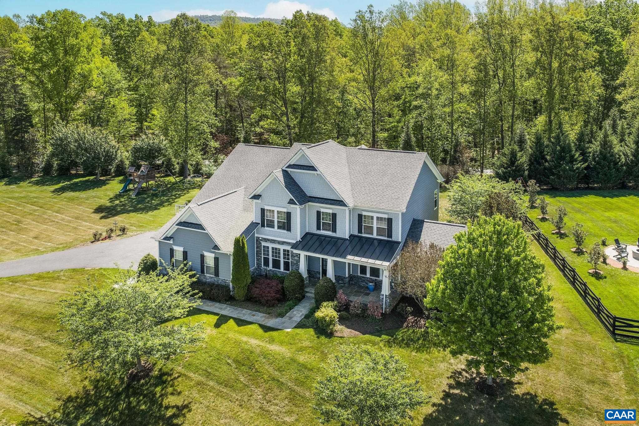 Aerial view of a gray two-story home with a covered porch, spacious lawn, and wooded backdrop.