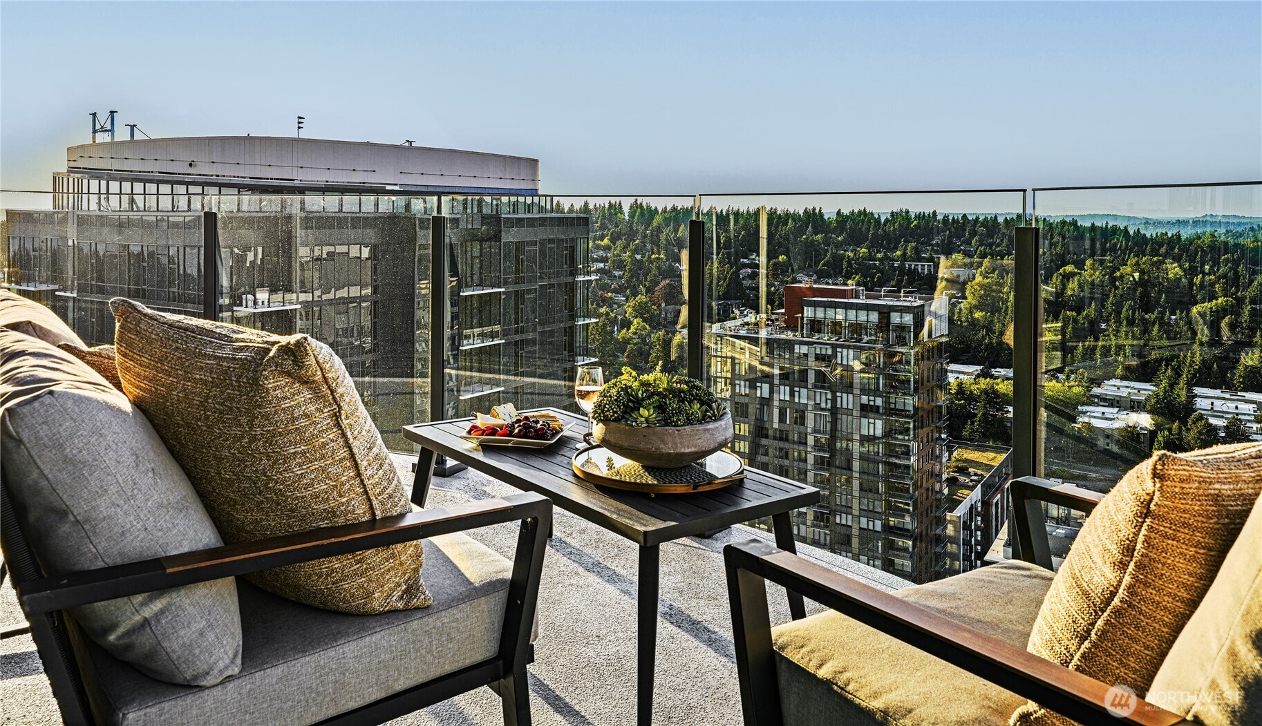 Furnished high-rise balcony with cushioned chairs, a coffee table, and views of buildings and forest.