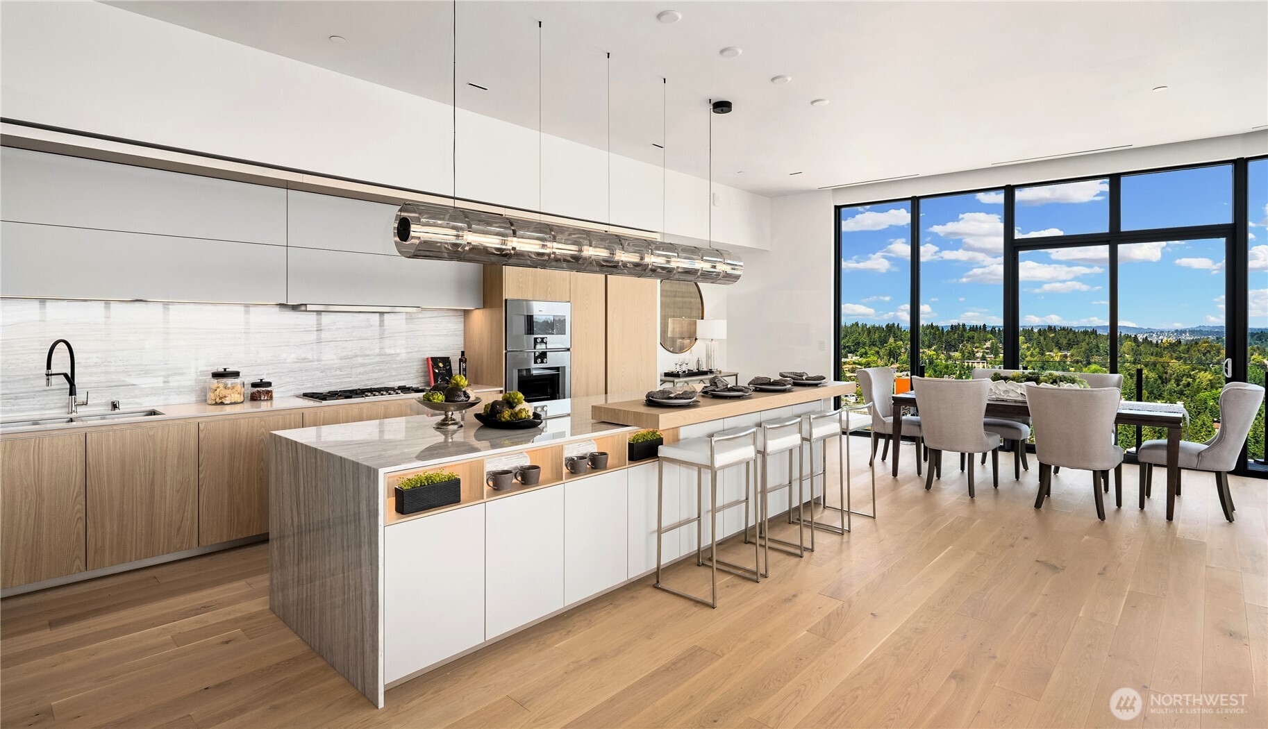 Modern kitchen with marble island, dining area, and expansive windows overlooking green treetops.