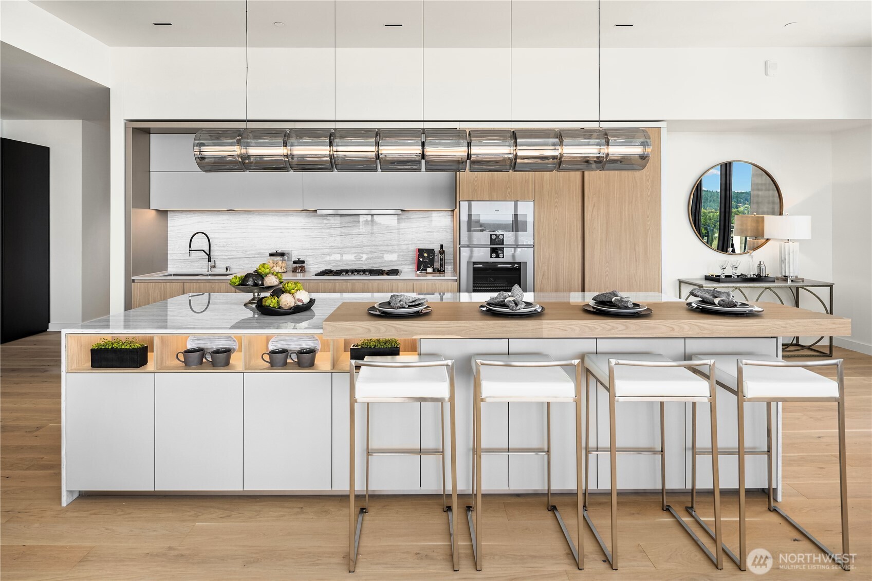 Modern kitchen with marble backsplash, light wood island, four white bar stools, and a sculptural glass pendant.