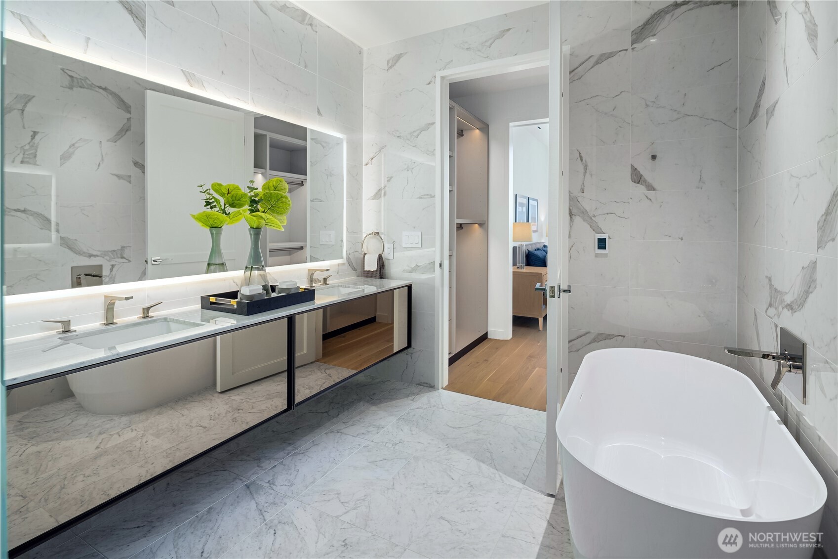 Marble-tiled bathroom with a long double vanity, illuminated mirror, and freestanding soaking tub.