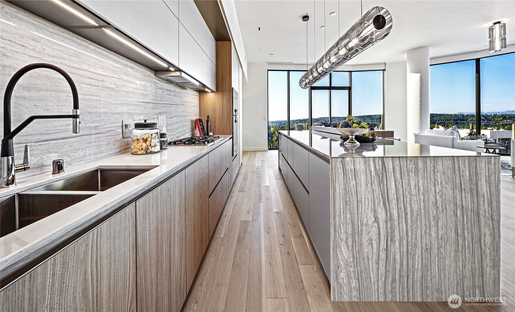 Modern kitchen with marble island and floor-to-ceiling windows overlooking wooded hills.