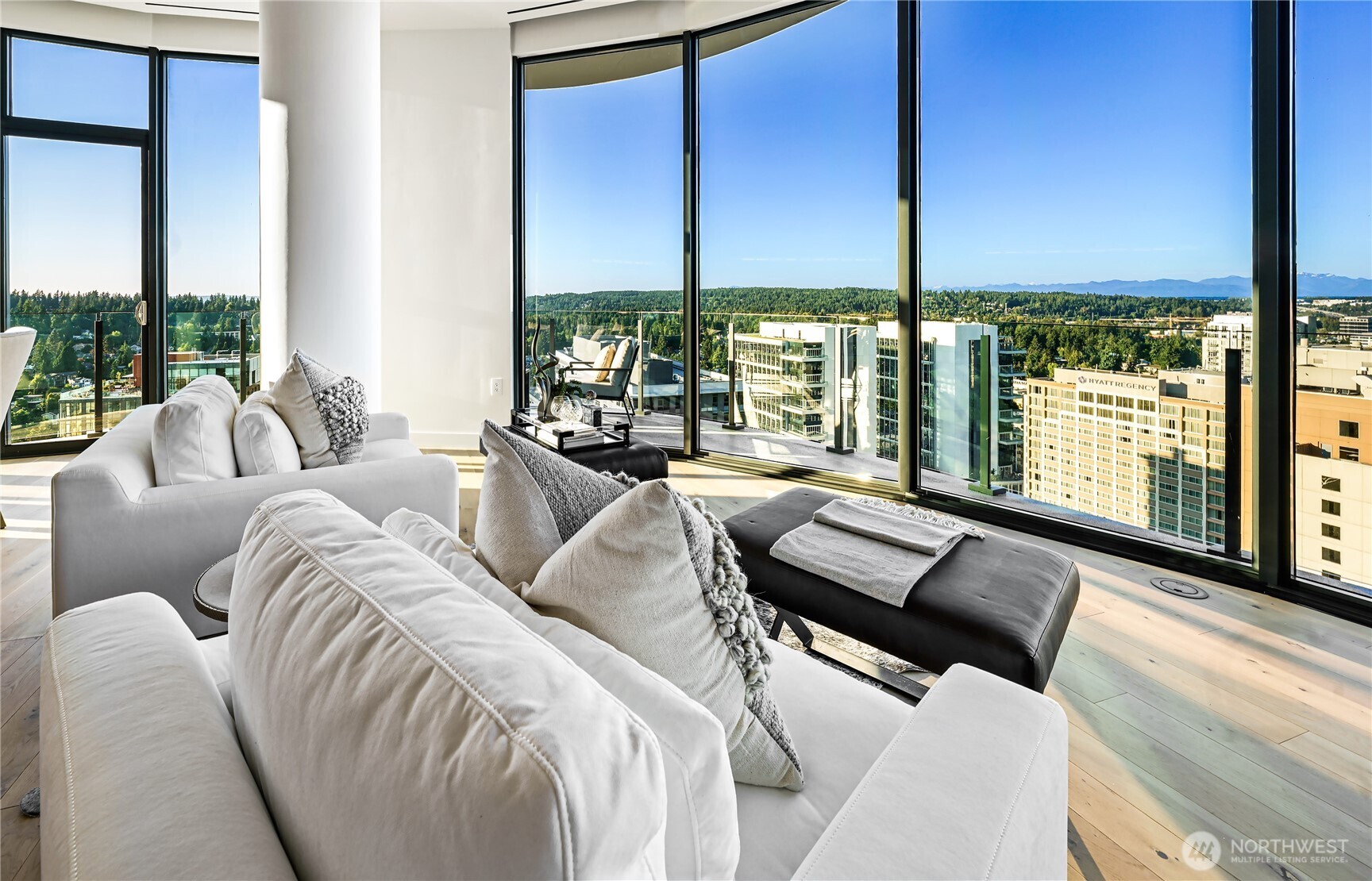 High-rise condo living room with white sofas, floor-to-ceiling windows, and Bellevue and mountain views.