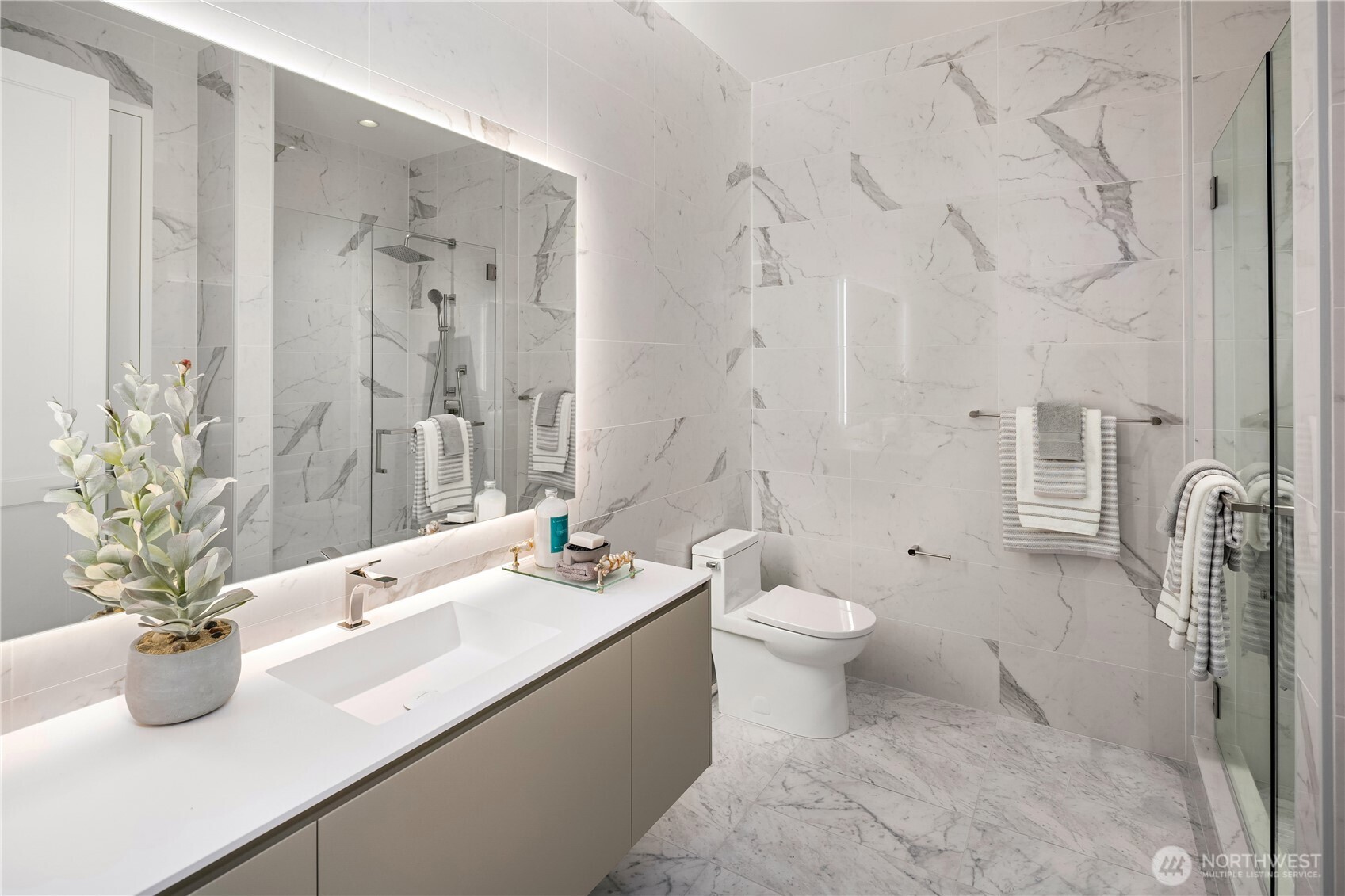 Marble-tiled bathroom with a wide vanity, illuminated mirror, glass shower, and toilet.