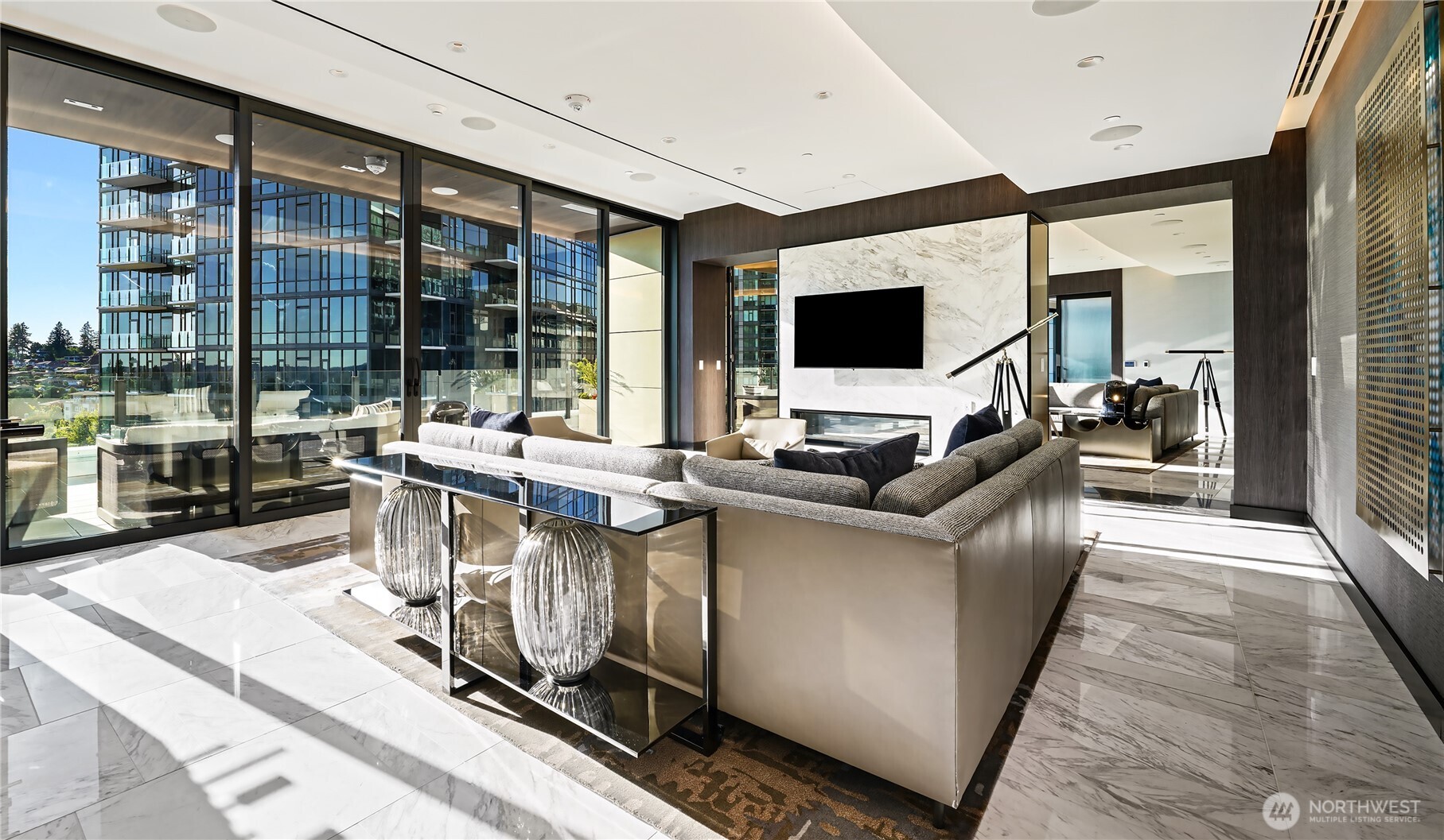 Furnished high-rise living room with a gray sectional, marble fireplace, and floor-to-ceiling windows overlooking glass towers.