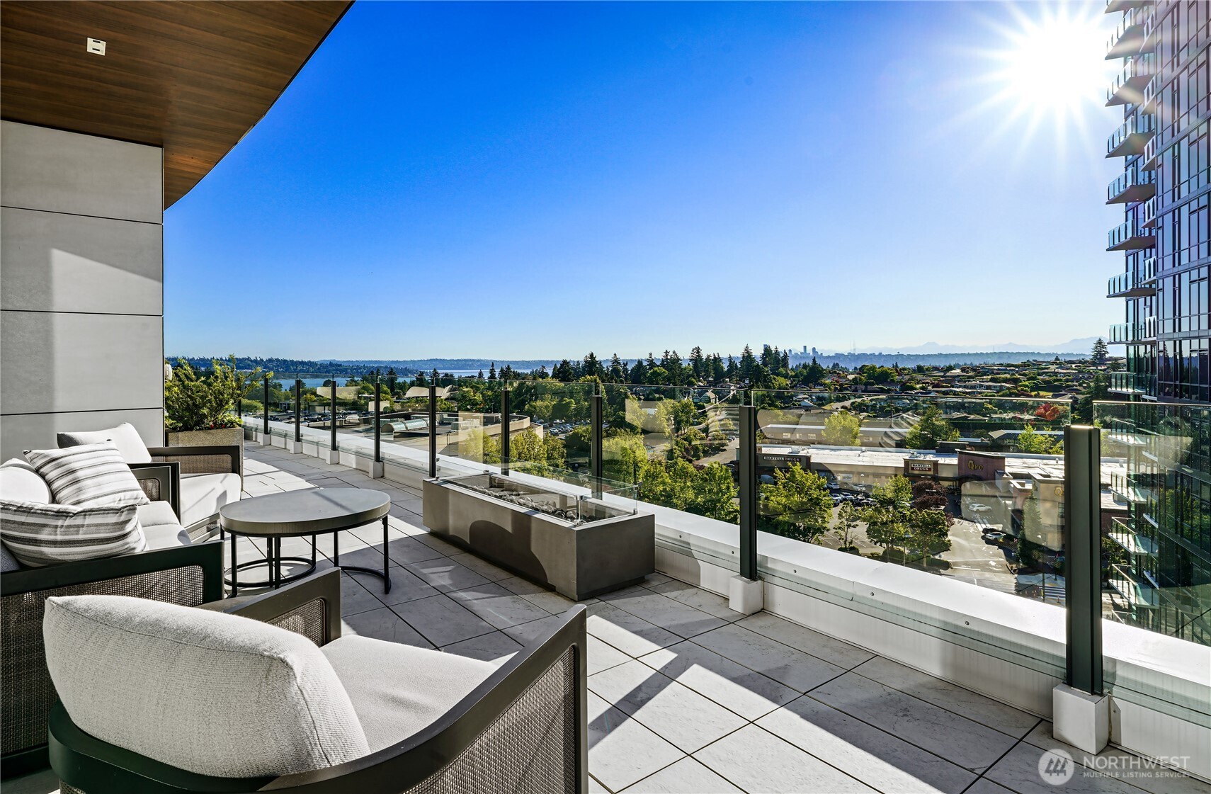 Furnished high-rise balcony with glass railing and sunny views of water, city, and distant mountains.