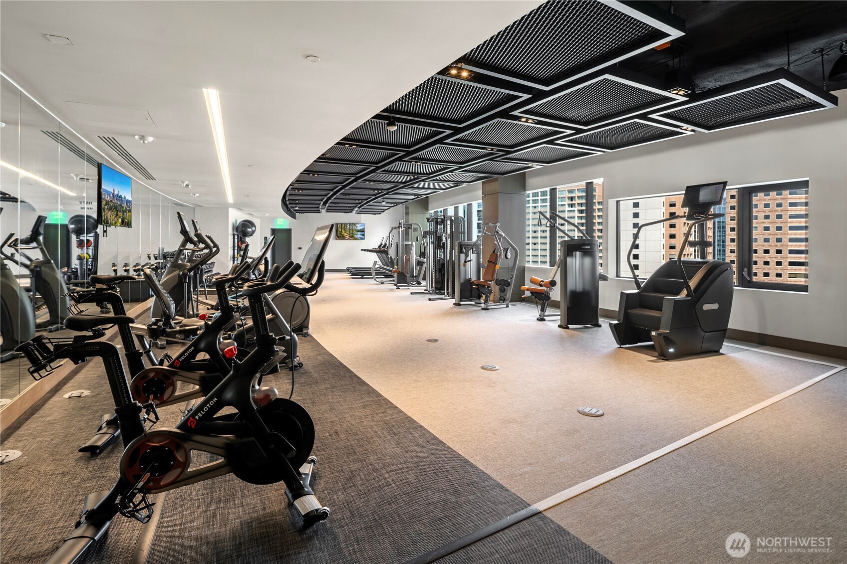 Condominium fitness center with Peloton bikes, strength machines, and large windows overlooking nearby buildings.