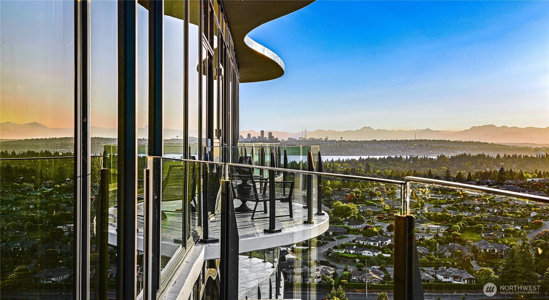 High-rise balcony with glass railings and seating overlooking the Seattle skyline and distant mountains at sunset.