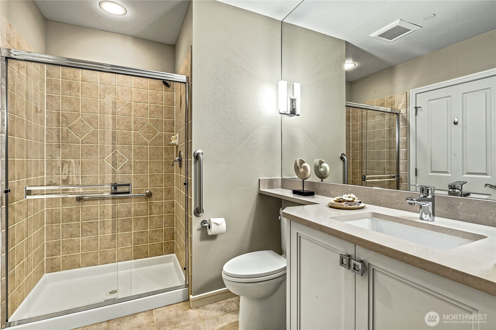 Bathroom with a tiled glass shower, toilet, and white vanity with a long mirrored countertop.