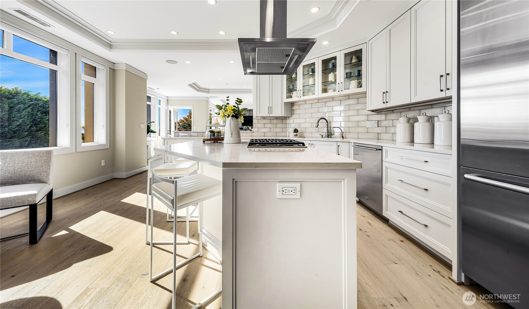 Bright white kitchen with a large cooktop island, glass-front cabinets, and broad windows.