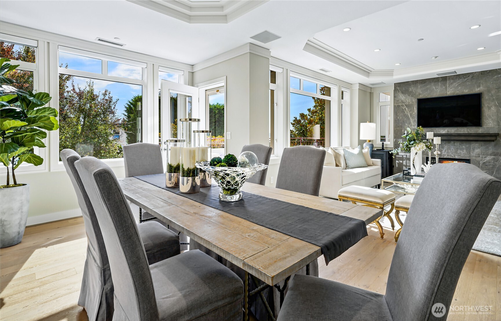 Dining area with a long wood table and gray chairs beside a bright living room and marble fireplace.