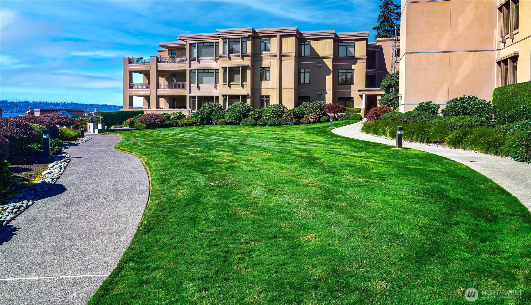 Condominium building beside a broad lawn and curved walkways, with landscaped gardens and distant water.