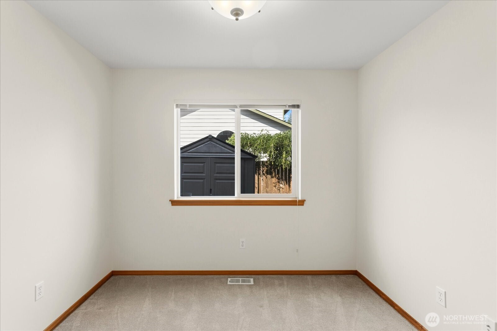 Empty carpeted room with a centered window, wood trim, and a view of a backyard shed.
