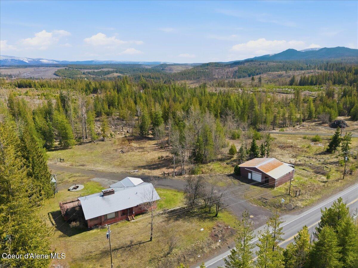 Rural property with a house and outbuilding beside a highway, backed by wooded hills and distant mountains.