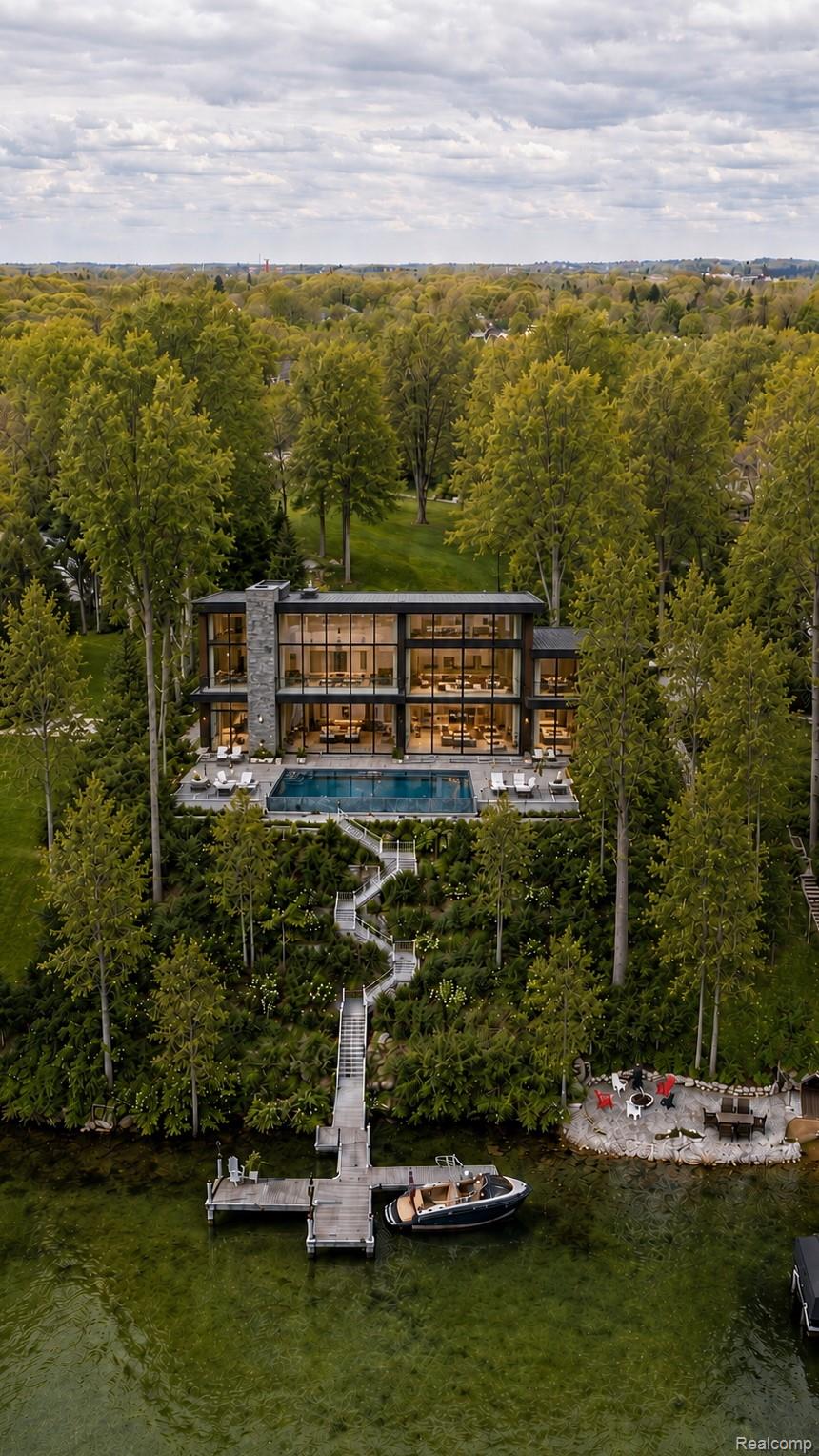 Modern lakefront home with a pool, long dock walkway, and boat among autumn trees.