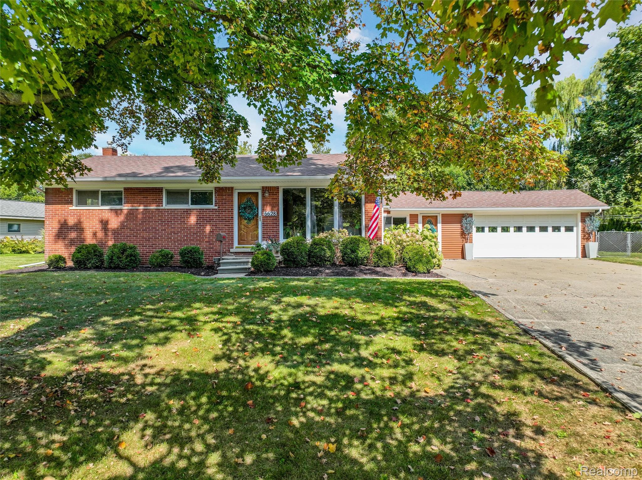 Red-brick ranch home with an attached garage, shaded lawn, and American flag.