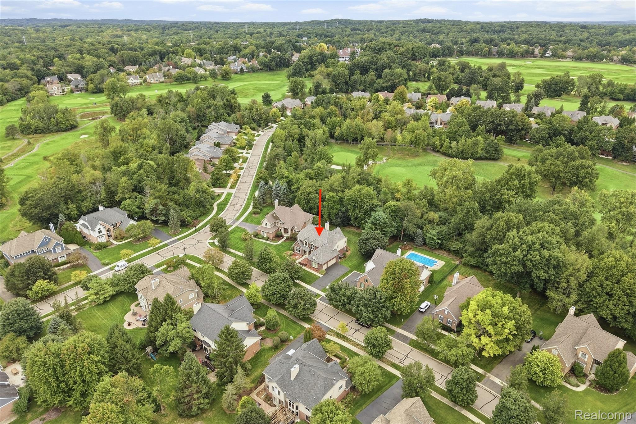 Aerial view of a two-story home marked by a red arrow, with a golf course and wooded neighborhood nearby.