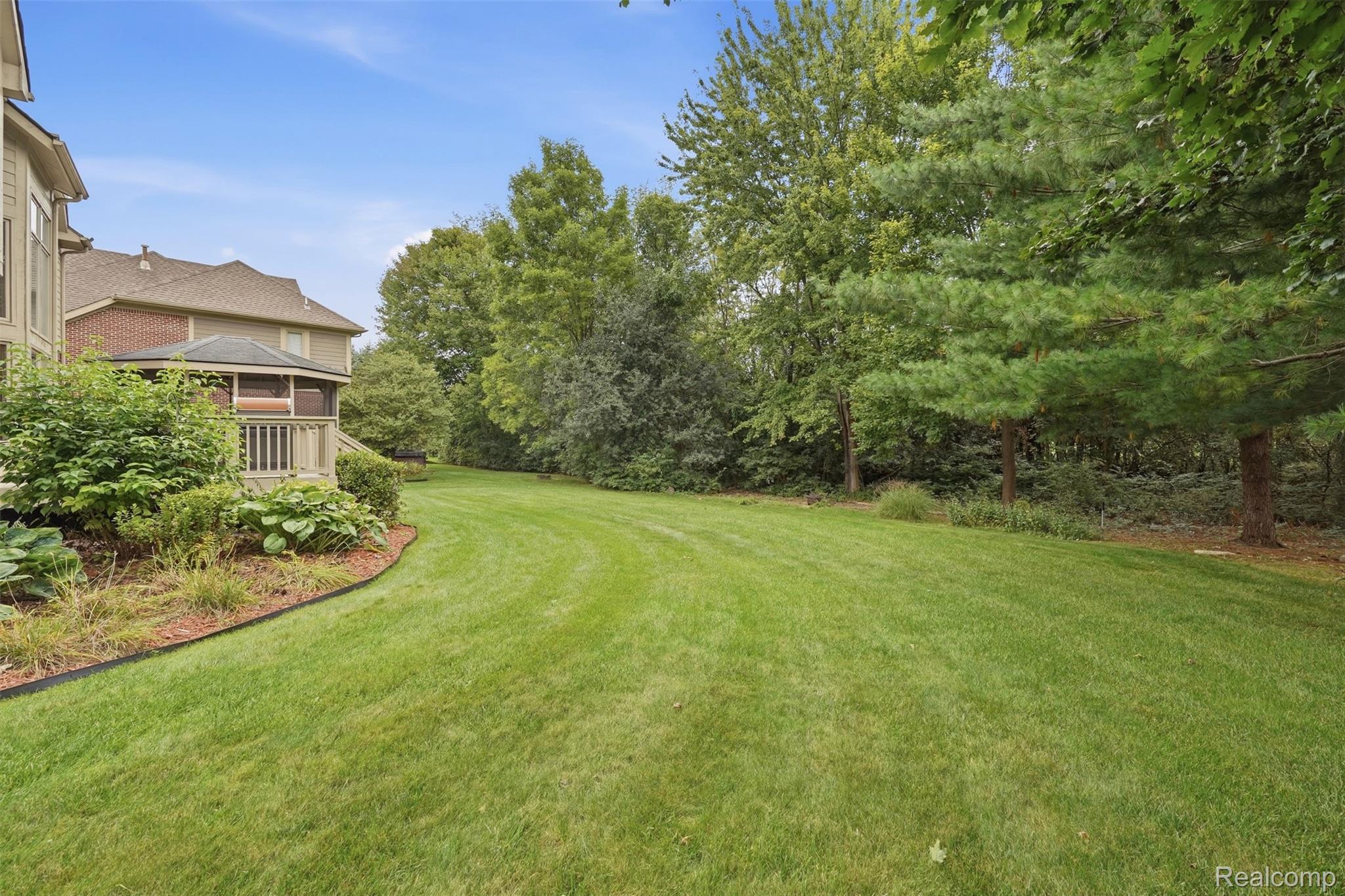 Spacious backyard lawn bordered by mature trees, with landscaped beds and a raised deck beside the home.
