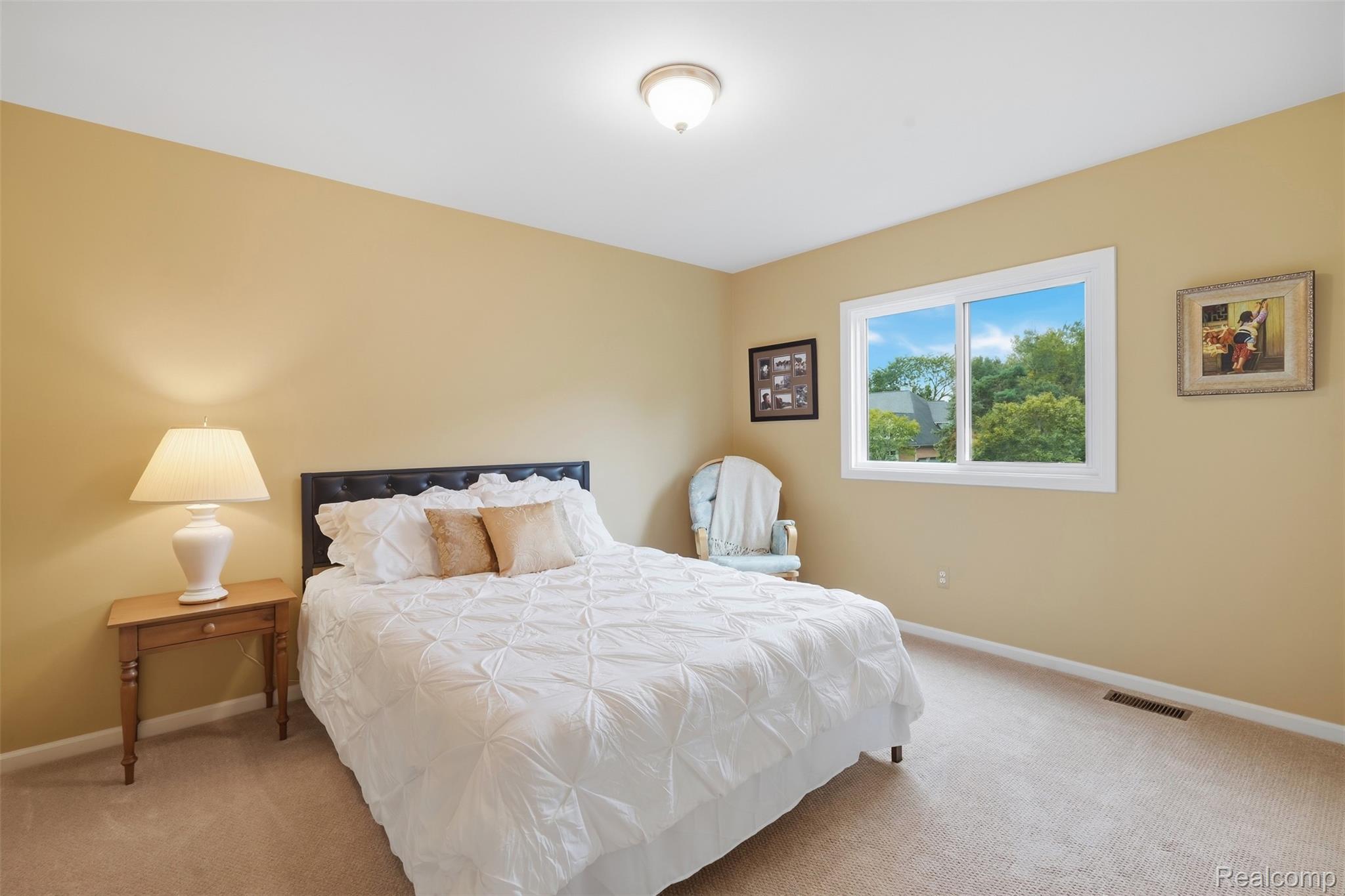 Bedroom with a white quilted bed, tan walls, bedside lamp, and window overlooking trees.