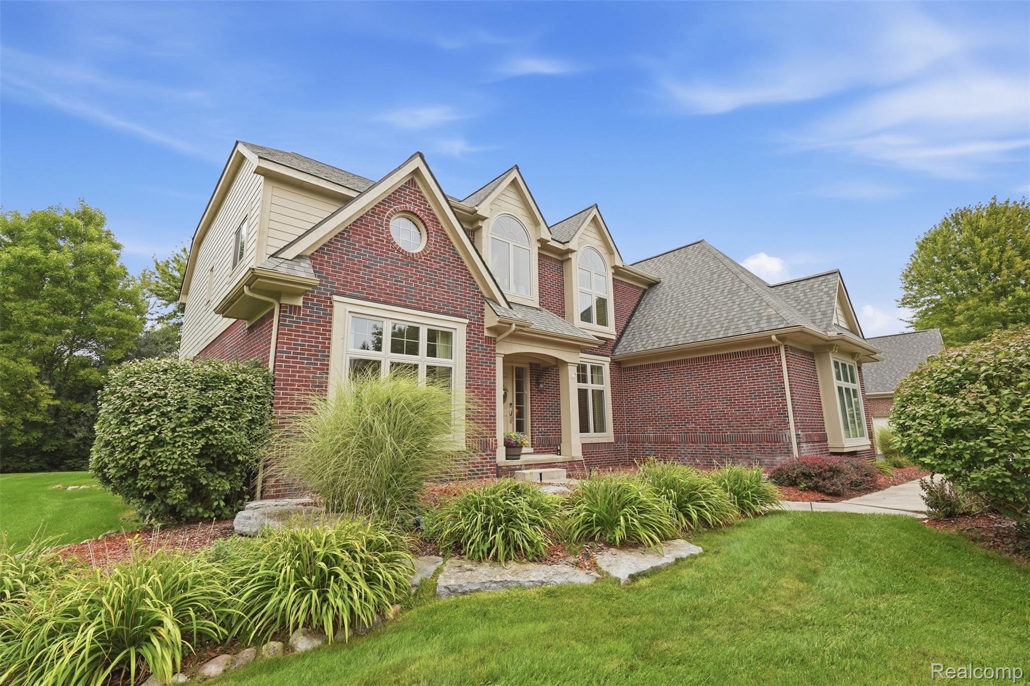 Two-story brick home with multiple gables, landscaped garden beds, and a broad green lawn.