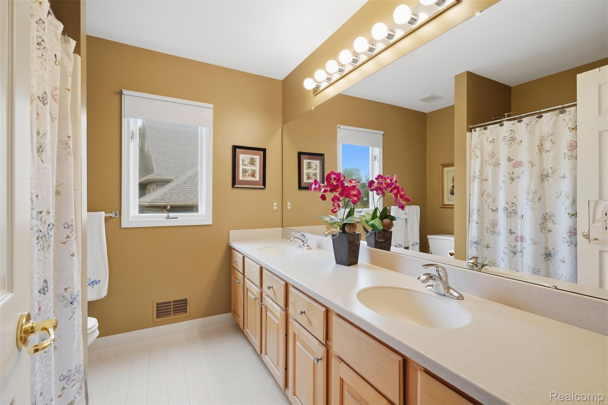 Bathroom with a double-sink vanity, large mirror, light wood cabinets, and floral shower curtains.