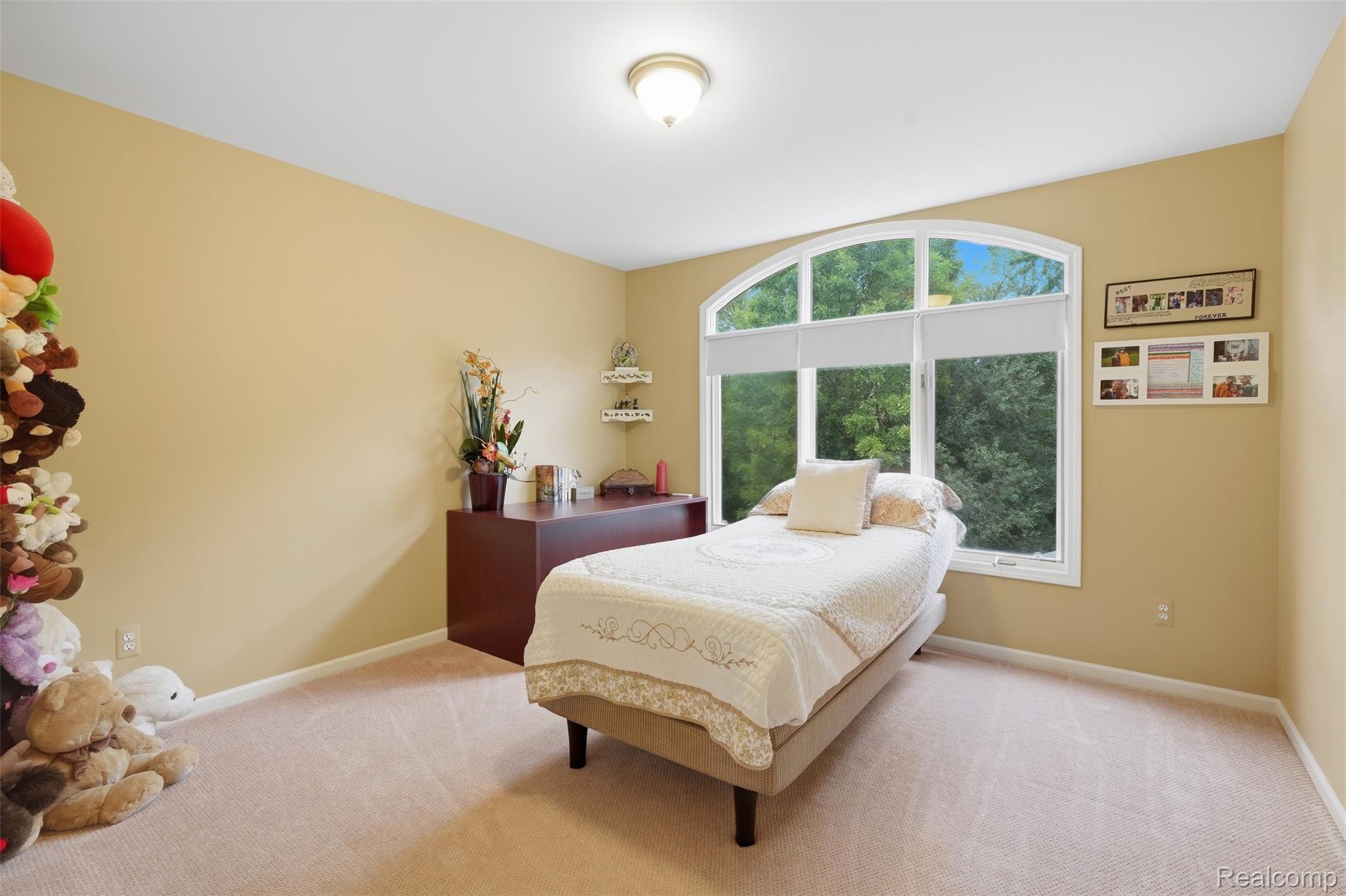 Bedroom with cream bedding, plush toys, and a large arched window overlooking trees.