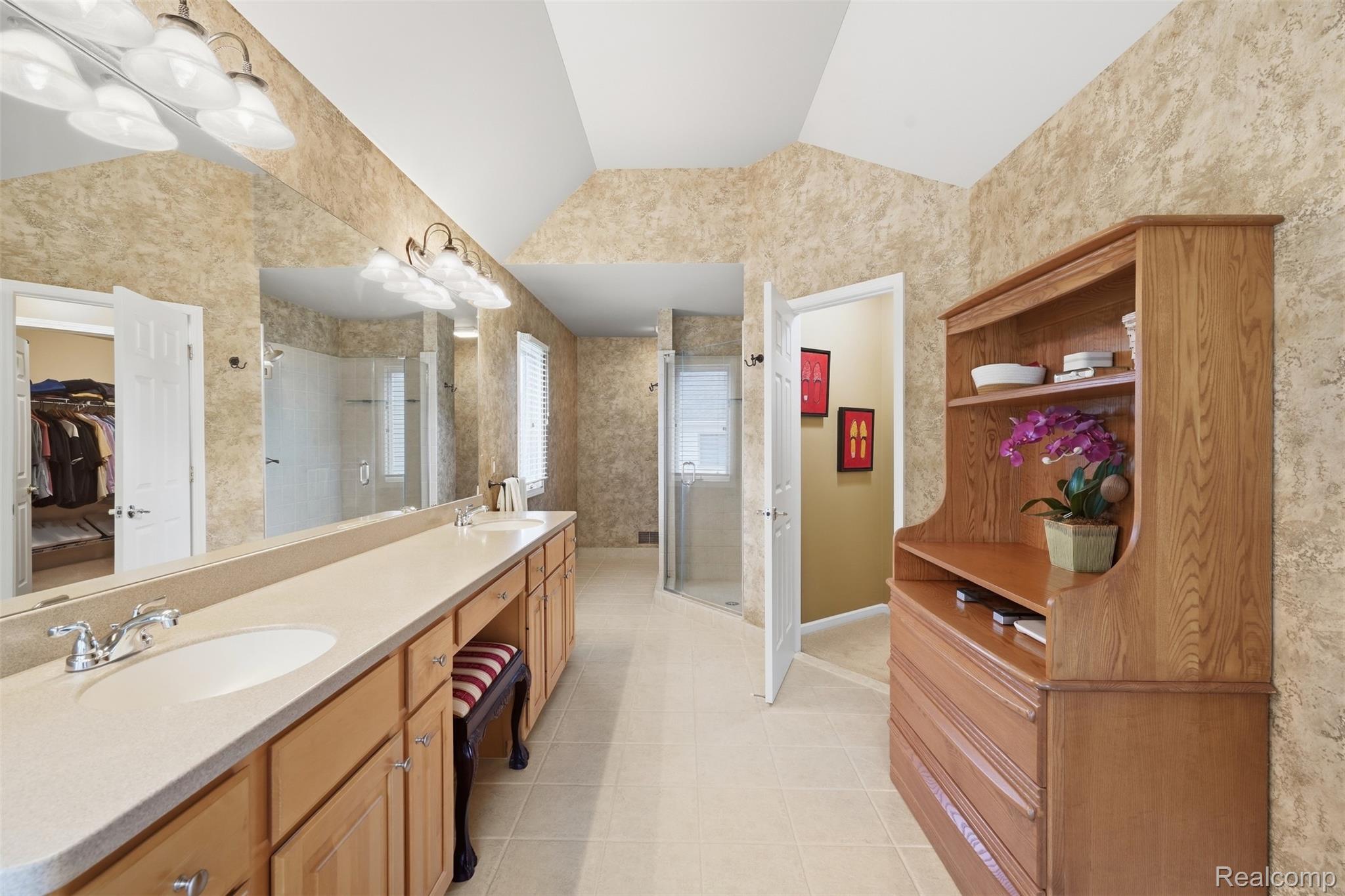 Primary bathroom with a long double vanity, glass shower, vaulted ceiling, and wooden built-in shelving.