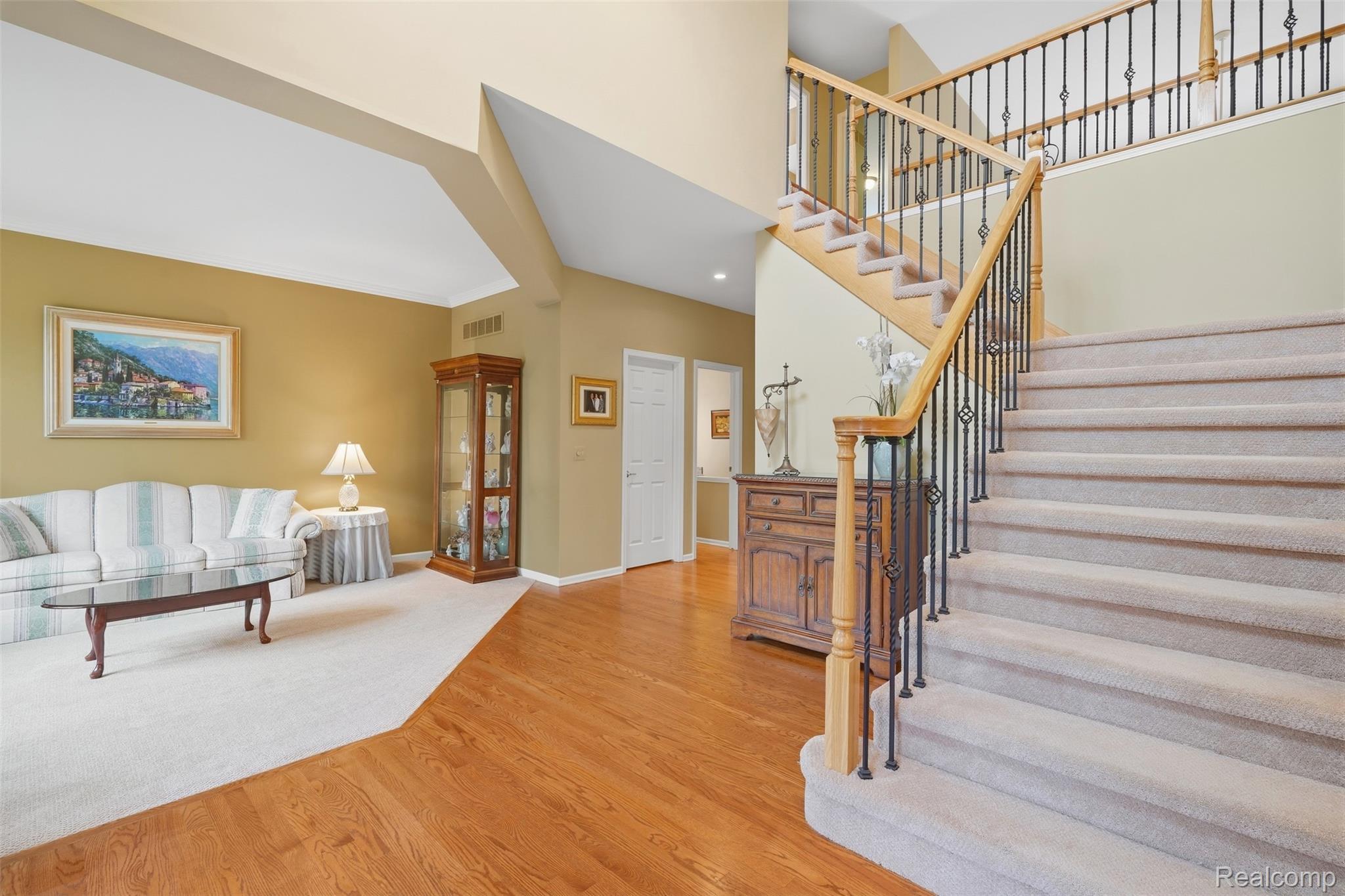 Furnished living room and entryway with a sweeping carpeted staircase and decorative black iron railings.