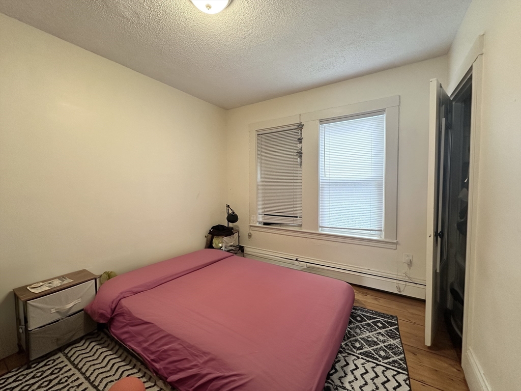 Bedroom with a pink-covered bed, two windows with blinds, hardwood floors, and a patterned rug.
