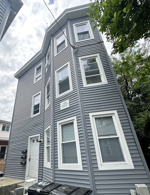 Tall gray-sided residence with white-trimmed windows, viewed from below beside leafy trees.