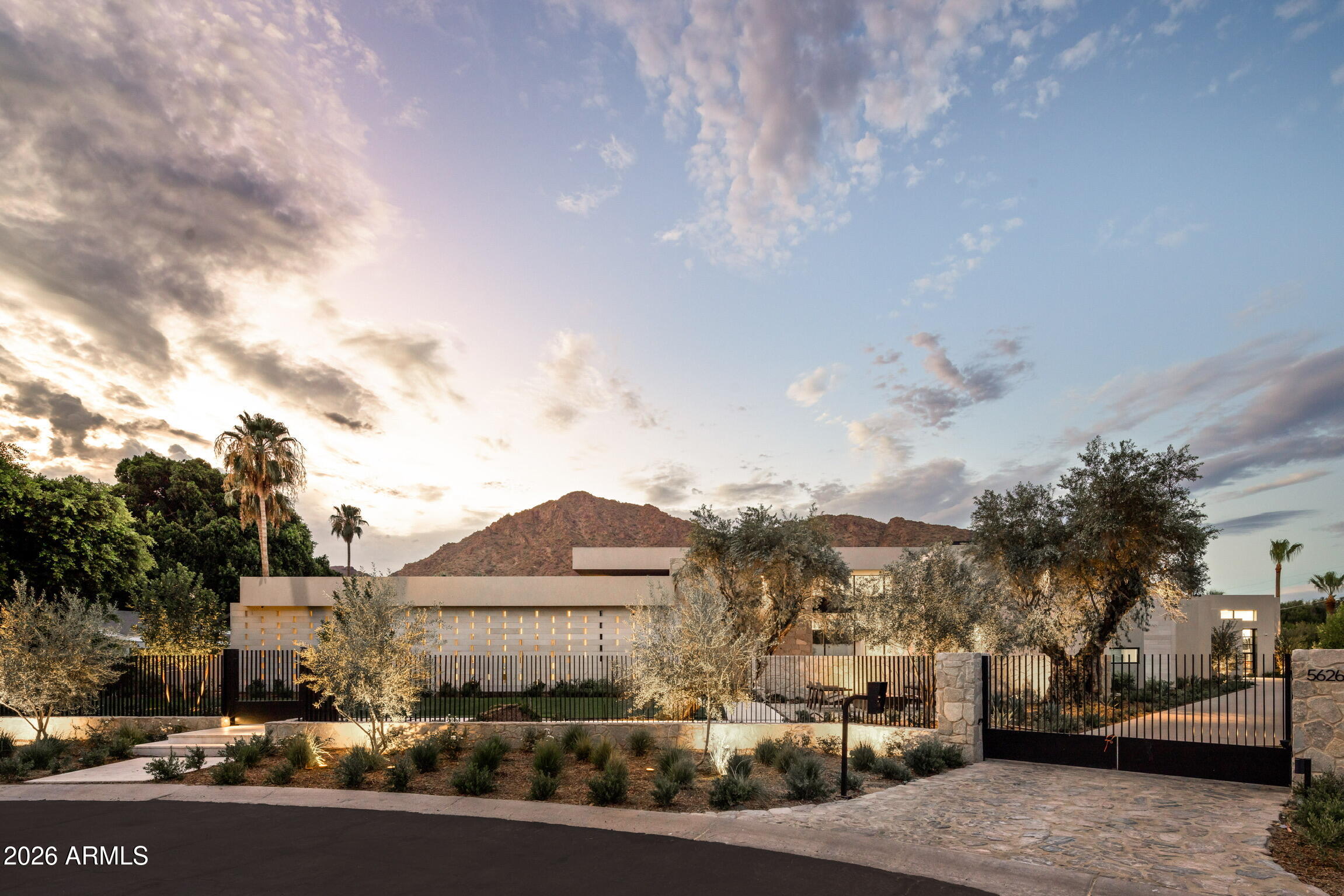 Modern gated home with desert landscaping and mountains behind it at sunset.