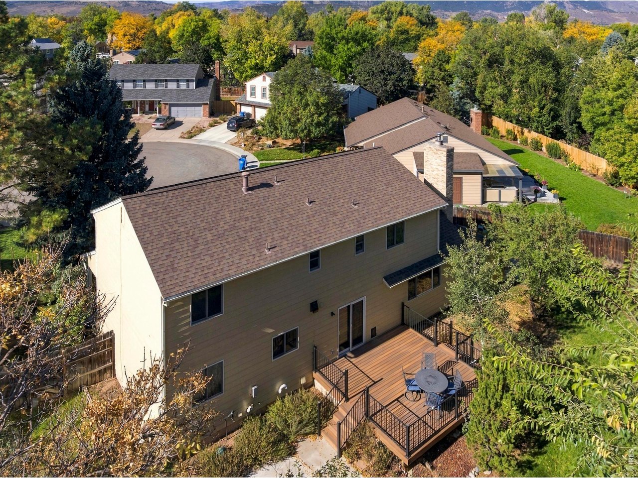 Aerial view of a two-story home with a large furnished deck, surrounded by autumn trees and distant foothills.