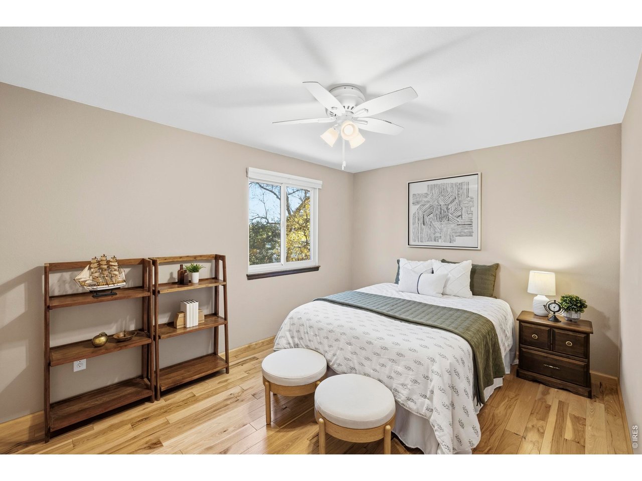 Staged bedroom with white and sage bedding, wooden shelves, light wood floors, and a window overlooking trees.