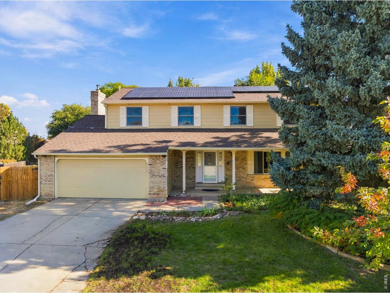 Two-story yellow-and-brick house with rooftop solar panels, attached garage, and a large evergreen tree.