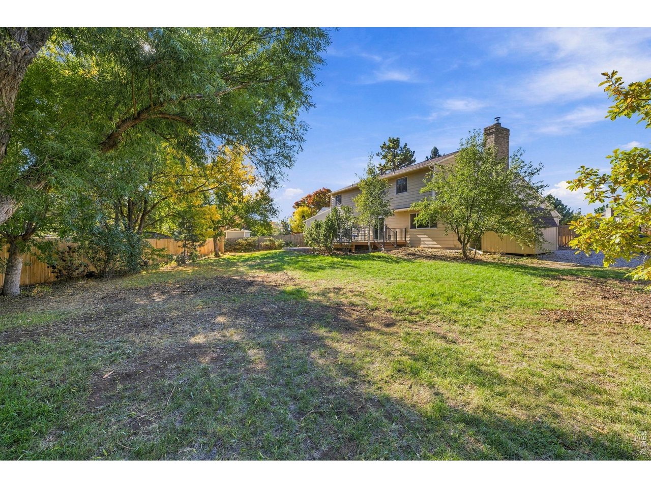 Two-story beige home and deck beyond a broad, tree-shaded backyard lawn.