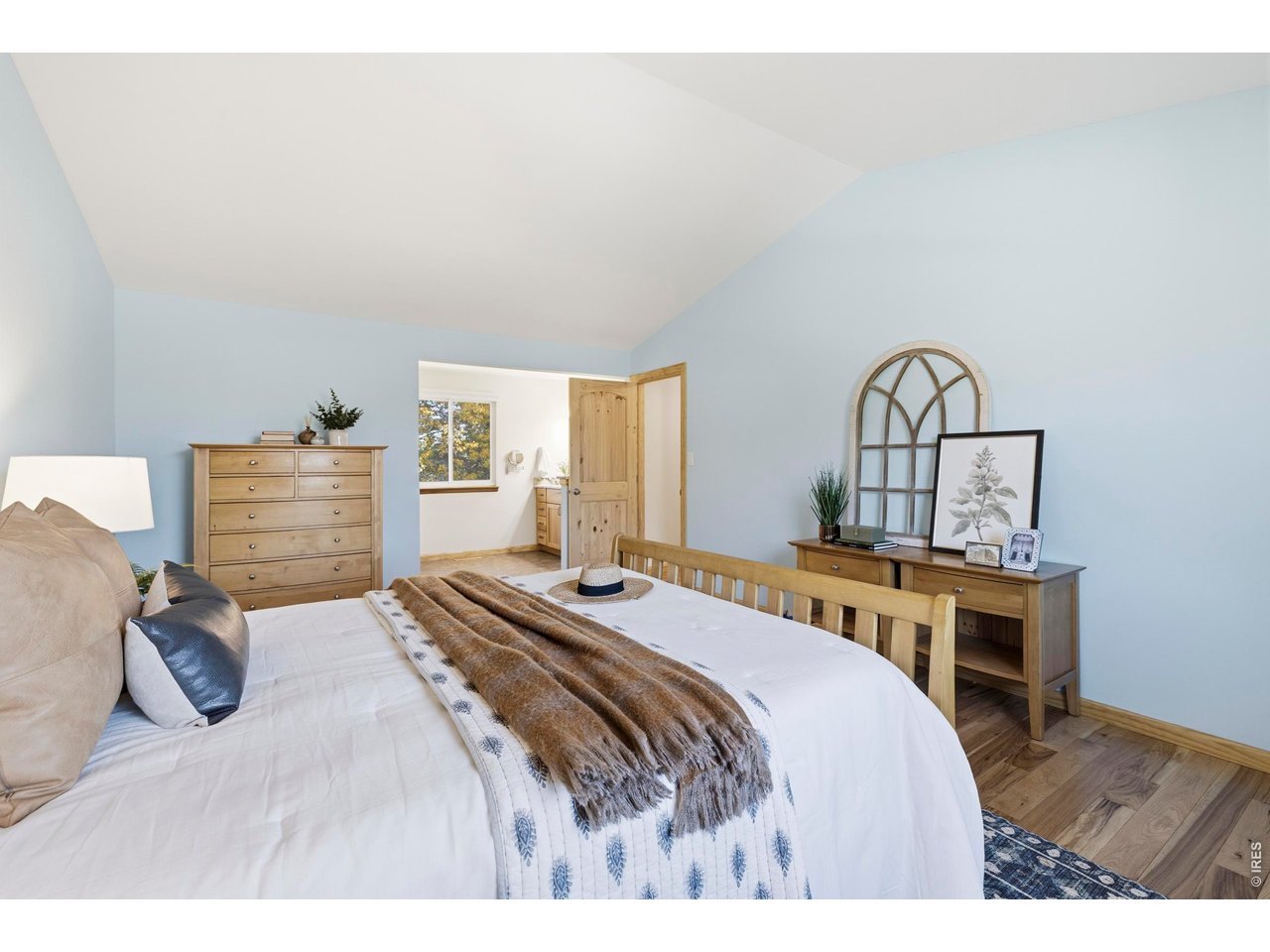 Furnished bedroom with a large bed, pale blue walls, vaulted ceiling, and light wood furniture.